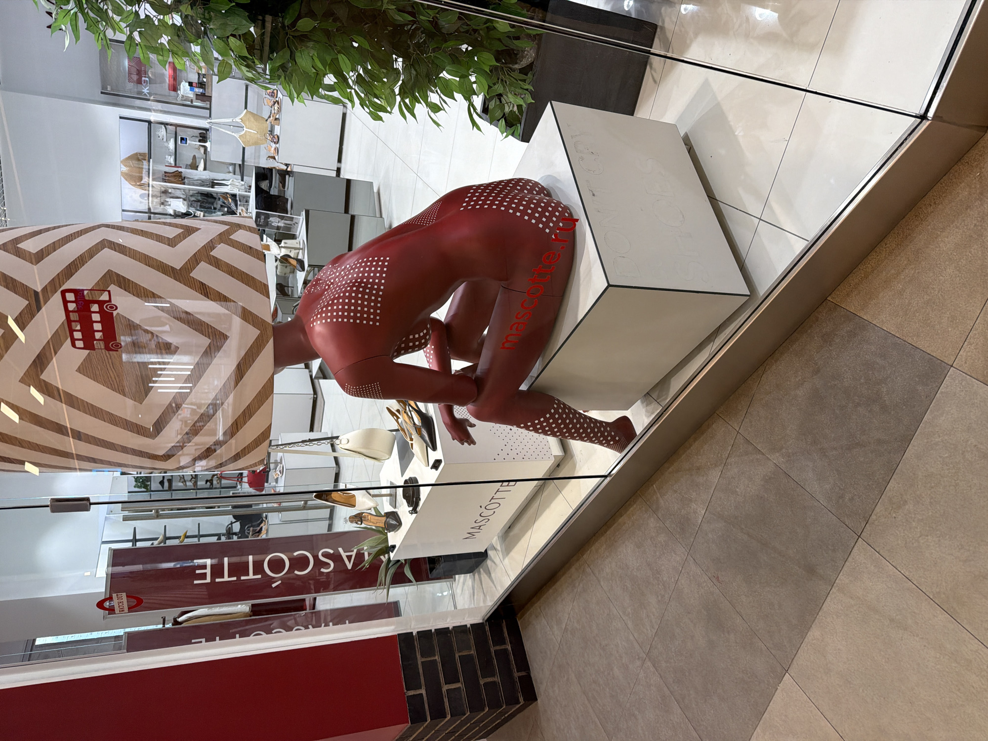 The photograph depicts a retail display inside a store, likely in Saint Petersburg, Russia, given the brand "MASCOTTE" and the Russian text on the mannequin. The display includes a red, dotted mannequin seated on a white block within a glass enclosure. Shoes and other accessories are arranged nearby. The store's interior is visible in the background, including shelving and other displays. A striped awning is partially visible above. The exterior flooring consists of large, square tiles.