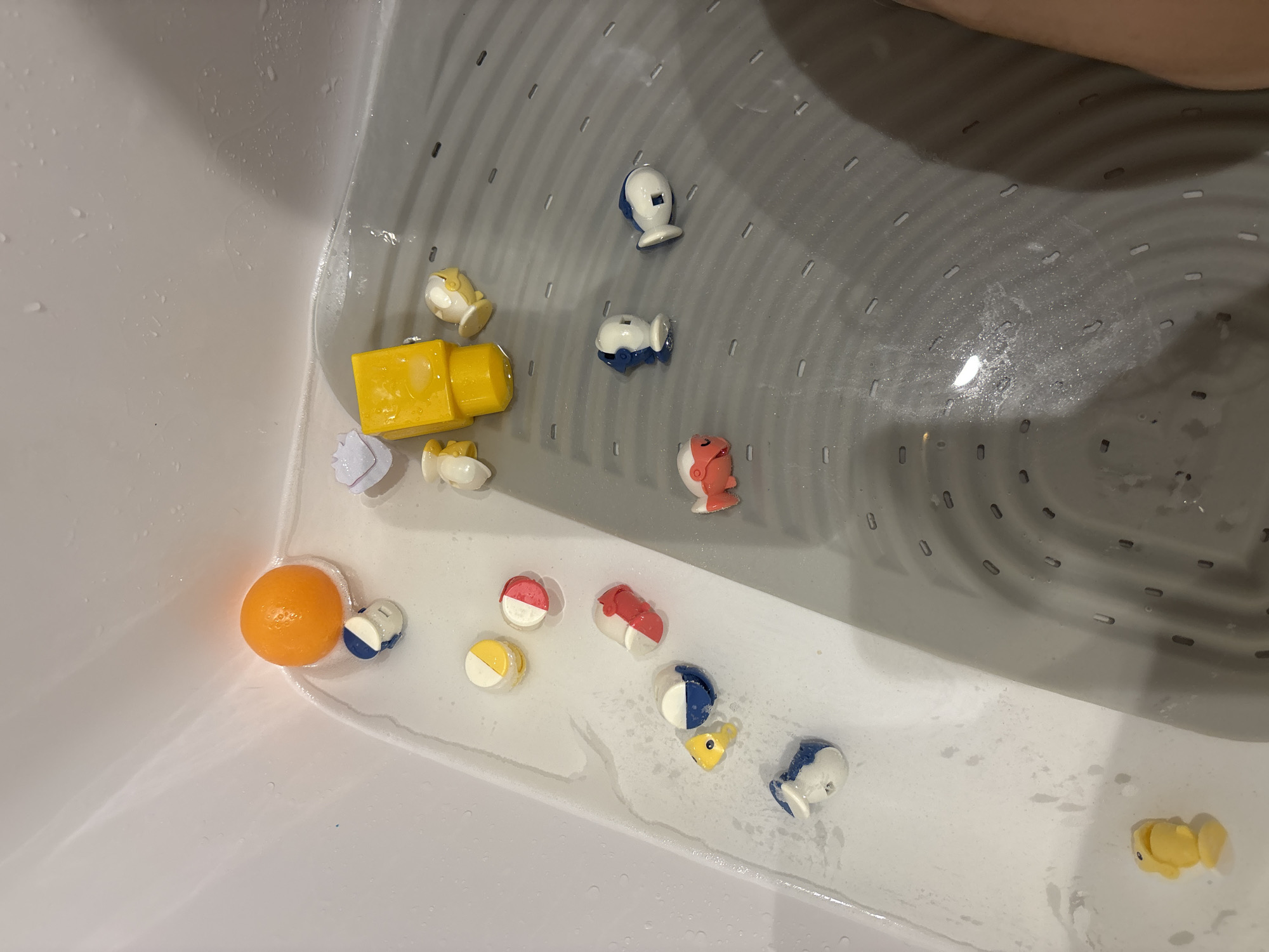 A close-up, overhead shot shows a white bathtub filled with soapy water and various bath toys. The toys include a bright orange ball, several small cylindrical toys with yellow and red accents, two white and blue fish-shaped toys, a red fish toy, a yellow duck, a yellow rectangular block, and a white shell-shaped toy. The bathtub has a gray textured bottom with a drain. The scene is indoors. No specific location in Dubai, United Arab Emirates is discernible from the image.