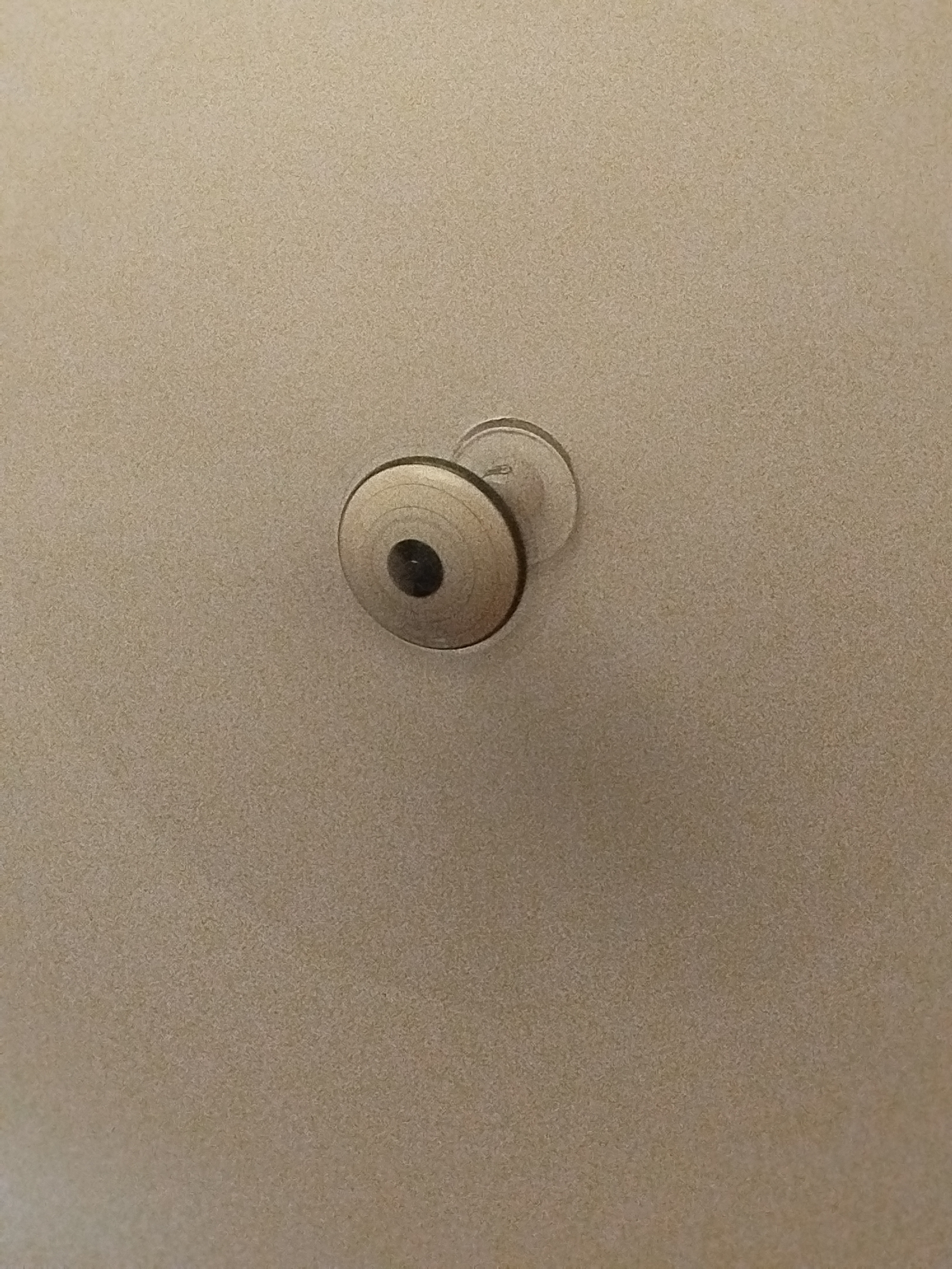 The image shows a close-up view of a light beige, textured surface, likely a ceiling. Centered within the frame is a round, metallic object attached to the surface. This object appears to be a ceiling light fixture or a decorative element. It has concentric rings and a dark, circular center. The lighting is subdued, casting a slight shadow around the object. There are no people or any other discernible objects or features in the image. The location in Lassan, Pakistan, is not visually indicated in the image.