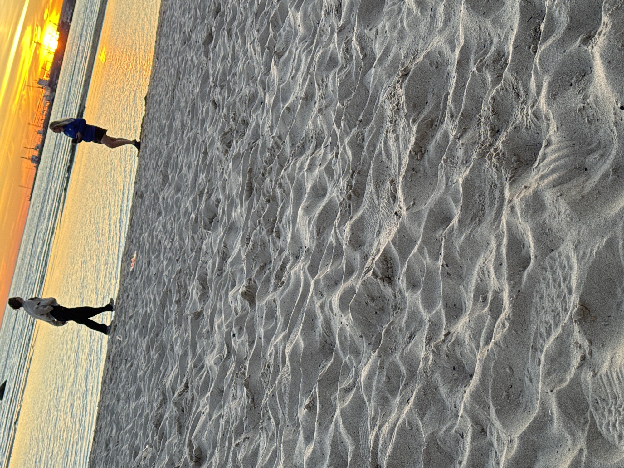 The image depicts a sandy beach at sunset, with the sea in the background. Two individuals are visible. On the left, a person wearing a light-colored jacket and dark pants walks along the shoreline. On the right, a person in a blue jacket and dark shorts stands near the water's edge, holding what appears to be a phone. The sky is a vibrant orange and yellow, indicating the sun is setting, with industrial structures visible on the horizon, suggestive of a port area. The foreground is dominated by the textured sand dunes. This scene is consistent with a location like Pirita Beach in Tallinn, Estonia, known for its sandy coastline and proximity to industrial port facilities.