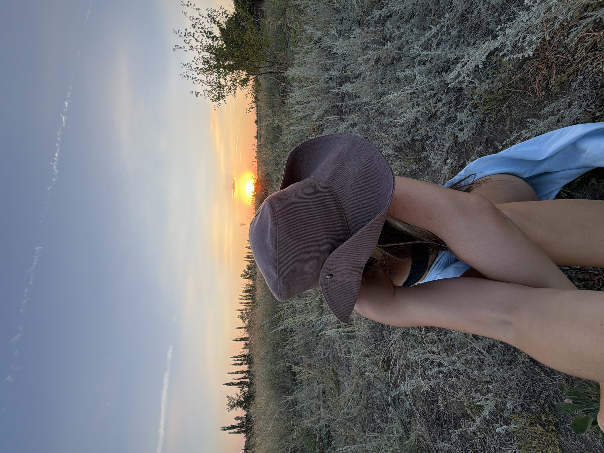 The image shows a person wearing a wide-brimmed hat and a light blue garment sitting in a field during sunset. The person is facing away from the camera, with their arms crossed in front of them. The sunset is visible in the background, with the sun low on the horizon, casting a warm glow over the scene. The field is covered in dry grass and some scattered bushes. In the distance, a line of trees is visible against the sky. The scene appears to be a peaceful and serene outdoor setting. The location is stated to be Nizhnyaya Lipovka, Russia.