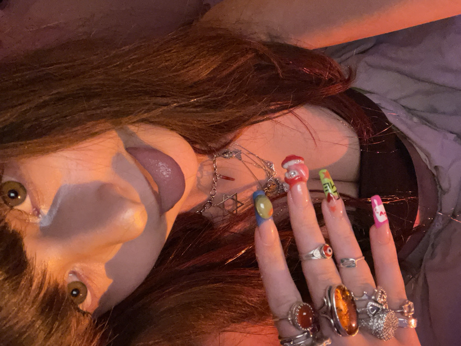 A person with hazel eyes and brown hair is shown from a low angle. Their mouth is slightly open, revealing their tongue. They are wearing necklaces, and their hand is prominently displayed with numerous rings. Each fingernail is painted differently, featuring designs such as a mushroom, words, and various colors. The background consists of soft, muted colors, with the person seemingly lying on a bed or soft surface.