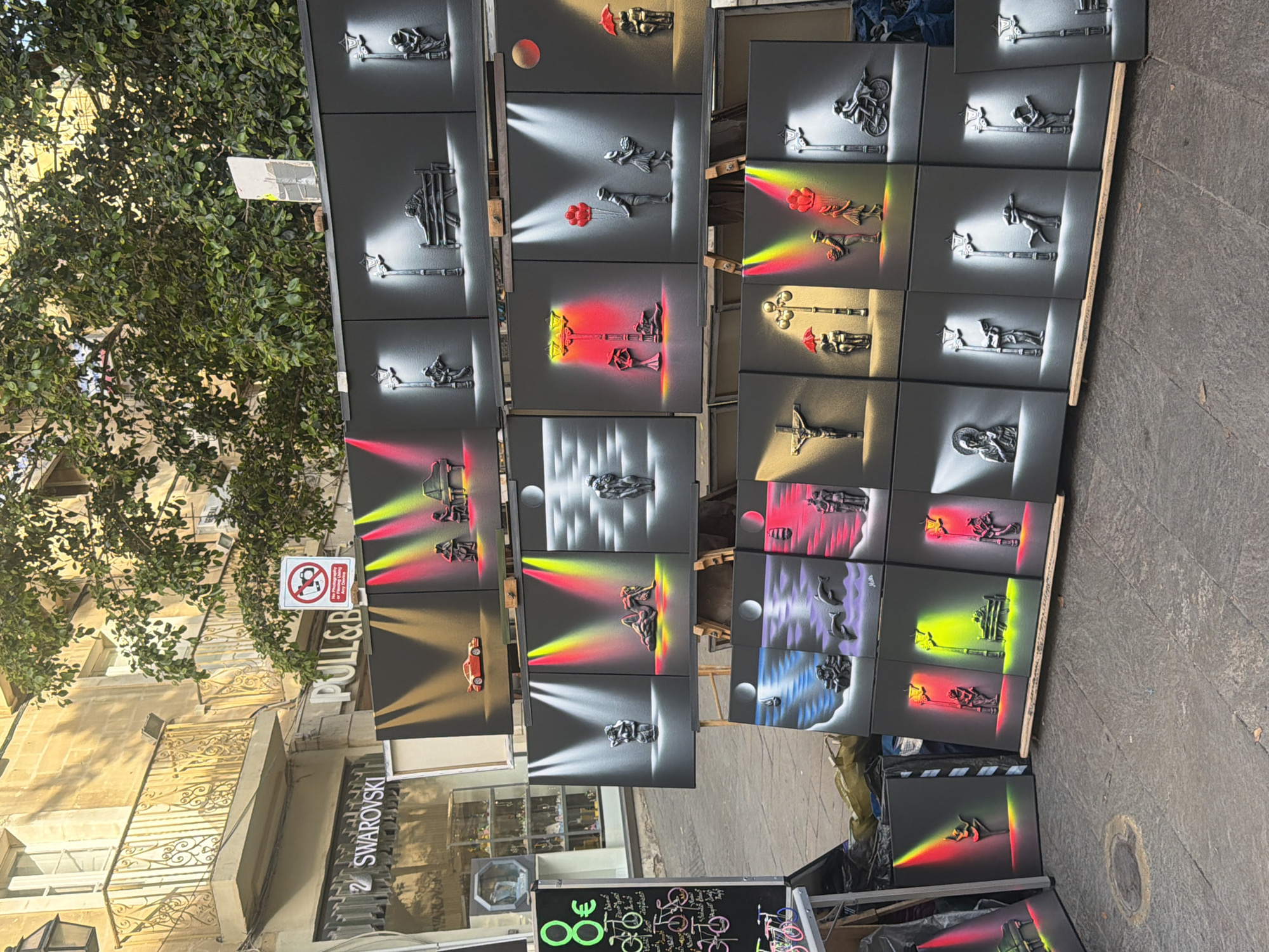 A vendor displays street art paintings on an easel in the city of Valletta, Malta. The paintings depict gray figures and objects against black backgrounds illuminated by brightly colored spotlights. The vendor's setup sits on a paved sidewalk. In the background are buildings, including a shop with a "SWAROVSKI" sign, trees, and a street sign.