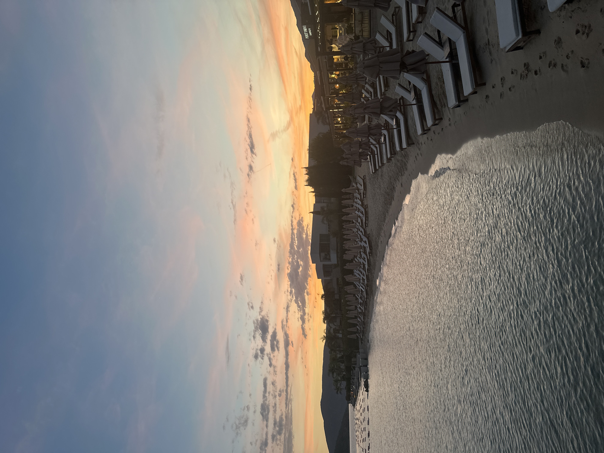 The image captures a coastal scene at sunset in Herceg Novi, Montenegro, specifically at the Portonovi resort. The sky transitions from pale blue at the top to vibrant orange, pink, and yellow tones near the horizon, interspersed with scattered grey and white clouds. The foreground and midground on the left consist of calm, dark water with visible ripples, where numerous dark, rectangular buoys or platforms are arranged in parallel lines. A light-colored sandy beach curves from the left midground towards the right foreground, densely populated with multiple rows of unoccupied sun loungers, each accompanied by a closed, light-toned beach umbrella. Footprints are visible in the sand near the waterline. Behind the beach, a modern resort complex with light-colored buildings and large windows extends along the shore, partially obscured by palm trees and other foliage. On the far right, a commercial structure with illuminated "PORTONOVI FOOD & B" signage is prominent. Dark, undulating mountains form the distant left background, silhouetted against the sky. No individuals are present in the scene.