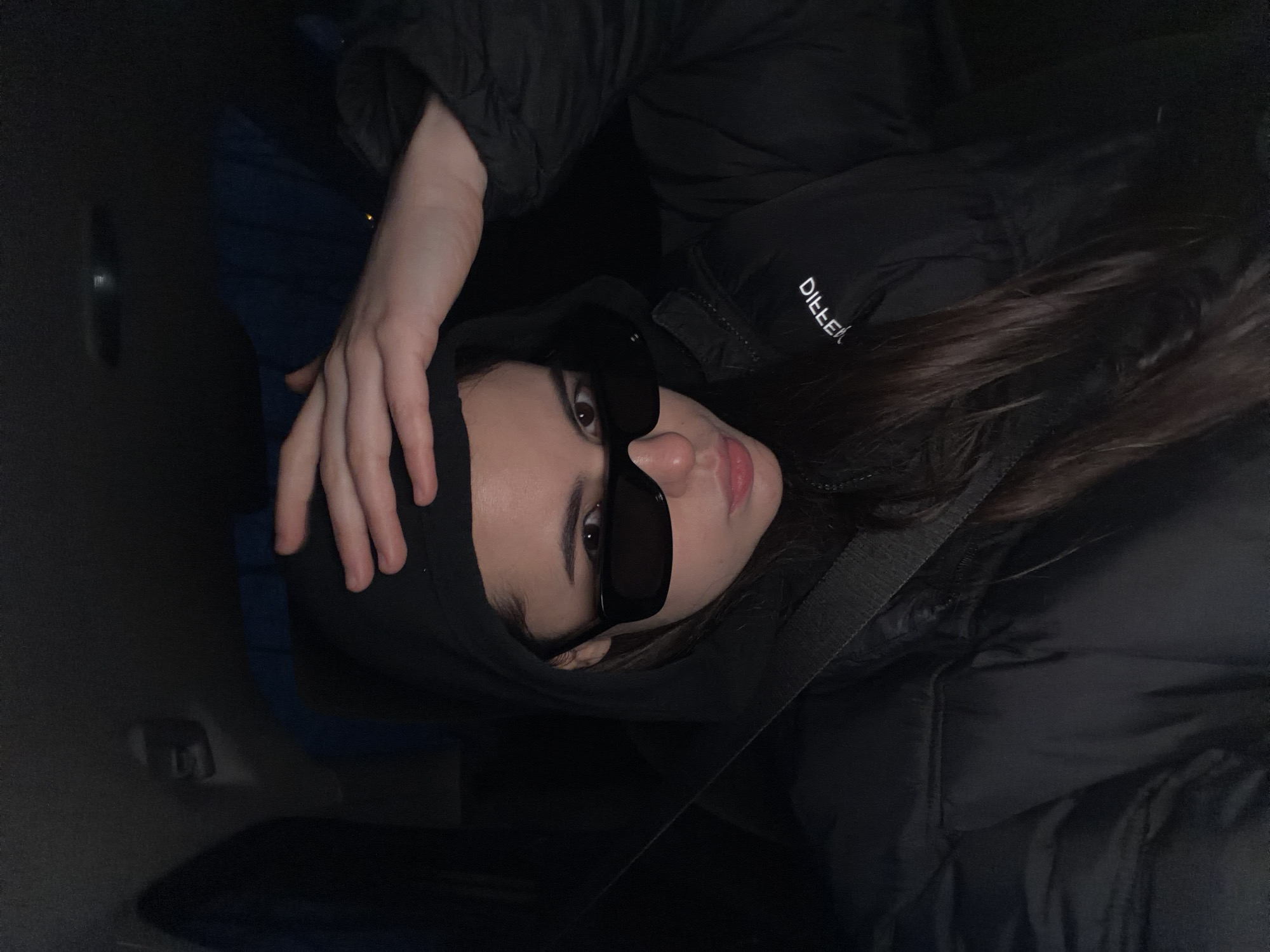 The image features a young individual, likely female, seated in a car, possibly in the passenger seat, in Chaikovsky, Russia. The subject wears a black hooded puffer jacket and black, rectangular sunglasses that obscure their eyes. Their dark hair is visible, and one hand is casually placed on the hood of their jacket, covering part of their head. A seatbelt is visible across their torso.
The car's interior is dimly lit, suggesting a nighttime setting or a poorly lit environment. Dark blue and black tones are discernible in the car's upholstery and interior panels. On the jacket, the white text "DIELLE" is visible, appearing upside down due to the image's original orientation. The heavy puffer jacket implies cool or cold weather. The person looks directly towards the viewer.