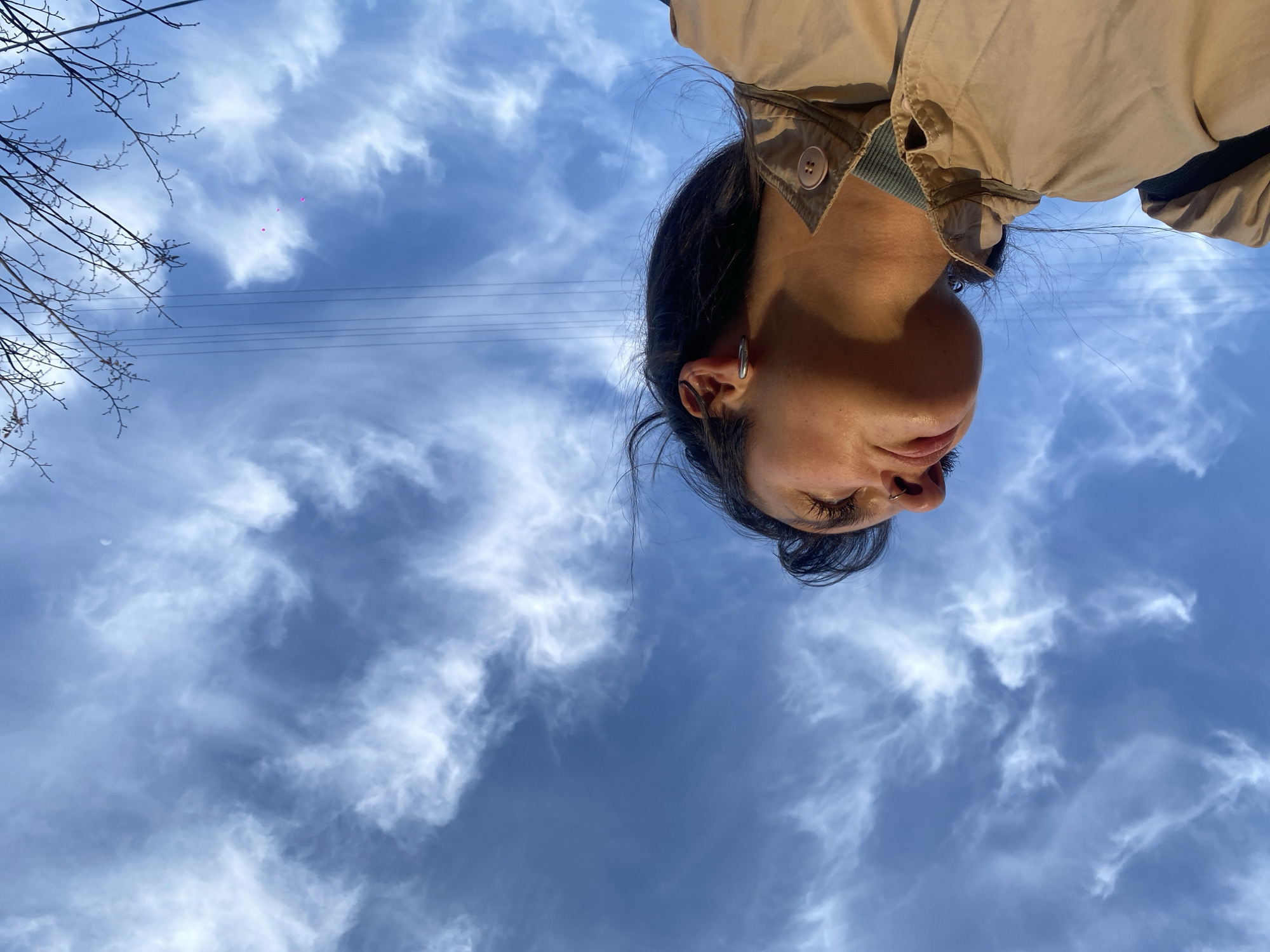 The image displays an upward-angled view of an individual positioned against a blue sky with cirrus clouds. The subject's face, visible from the chin upwards, is located in the lower right quadrant, illuminated by warm, directional light. The person has dark hair, a closed or squinted right eye with visible eyelashes, and a silver hoop piercing in the right nostril. A silver-colored hoop earring is present on the right earlobe. The individual wears a light brown or tan collared jacket with a single visible button on the collar flap, over a ribbed, possibly green or blue-green, inner layer. The sky occupies the majority of the frame, showing thin white clouds. In the upper left, multiple parallel utility lines are visible. Bare tree branches extend into the frame from the upper right corner. Three small, distinct magenta-colored objects, possibly balloons, are observed floating in the mid-left sky. The scene is outdoors in the city of Moscow, Russia.
