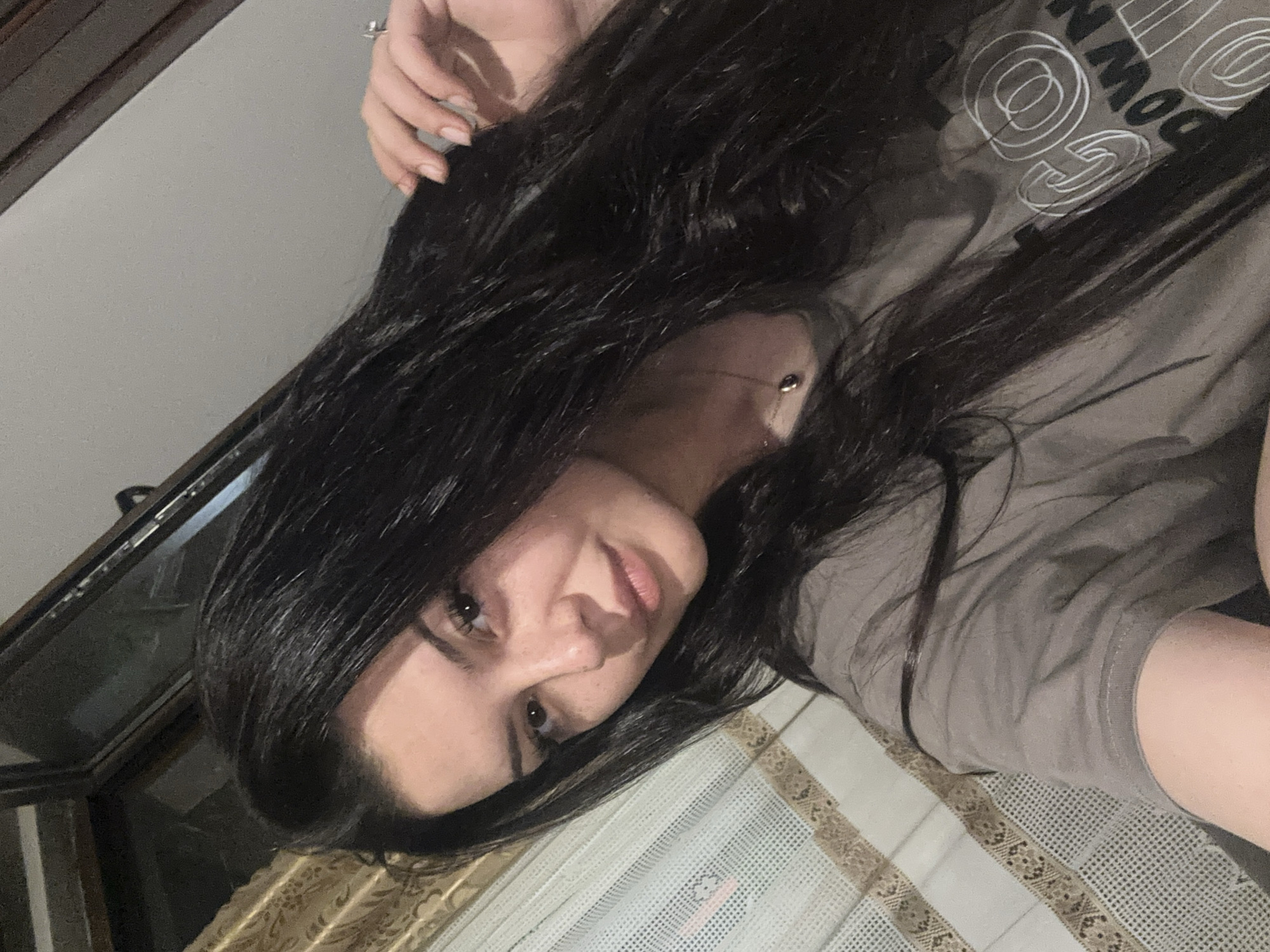 The original media file displays a young woman in a rotated, almost horizontal, perspective. She has long, dark, slightly wavy hair that flows around her head and shoulders. Her head is positioned on the left side of the frame, with her face turned upwards and looking towards the viewer, featuring dark eyes and a soft, gentle smile. She appears to be wearing light makeup, with defined eyebrows and natural-toned lips.
A delicate, possibly gold-toned, necklace with a small pendant is visible around her neck. Her right hand, adorned with a ring on her ring finger, is seen near the top of her head, lightly touching her hair. She is wearing a plain grey t-shirt that features white graphic text and circular designs. Partial words like "DOWN" and "GO" are visible on the fabric.
The background indicates an indoor setting, with a plain, light-colored surface (likely a wall or ceiling if the image were upright) and a patterned fabric, possibly a curtain or tablecloth, below her. There is also a dark, reflective surface, which might be a window or mirror frame, to the left of her head. The lighting is soft and even, suggesting an indoor environment without harsh direct light. The image captures a casual, relaxed moment, possibly a selfie, set within a home in Chubra, Bulgaria.