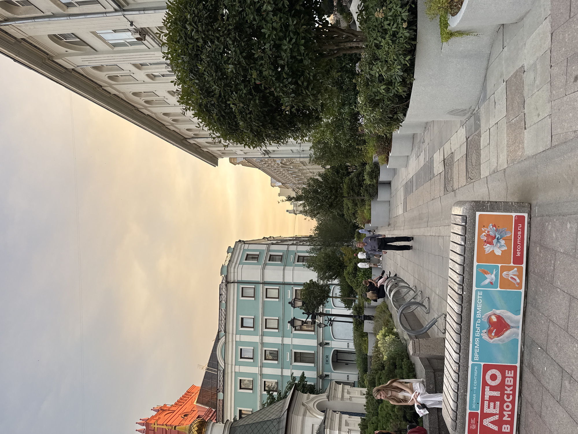 The image depicts a street scene in Moscow, Russia, likely during the evening or late afternoon as suggested by the warm, golden light in the sky. The perspective is from a pedestrian walkway with a view of buildings and greenery.
On the left, a prominent building with a light blue facade and white trim dominates the mid-ground. Above it, a glimpse of a red-colored building with golden domes and a cross can be seen, hinting at a religious structure adjacent to the commercial area. A store with "DIOR" signage is visible on the ground floor of the blue building.
To the right, a large, white, multi-story building with arched windows stretches along the street. Lush green trees and shrubs are planted in modern, cylindrical planters along the sidewalk, creating a green buffer zone.
In the foreground, a concrete bench with wooden slats is prominently displayed. It features a large advertisement with Cyrillic text and colorful graphics, including images of a heart held by hands, a bird, and flowers. The text "ВРЕМЯ БЫТЬ ВМЕСТЕ" (Time to be together) is clearly visible.
Several people are present in the scene. A woman is seated on a low wall on the left, looking away from the camera. Further down the walkway, a group of people are seated and standing. A man in a grey shirt and dark pants is walking towards the viewer. Other individuals are scattered along the street, engaged in various activities like walking and sitting.
The ground is paved with stone tiles of various shades and sizes, with some manhole covers visible. Street lamps are also present, adding to the urban environment. The overall impression is a lively, contemporary urban space within the historic city of Moscow.