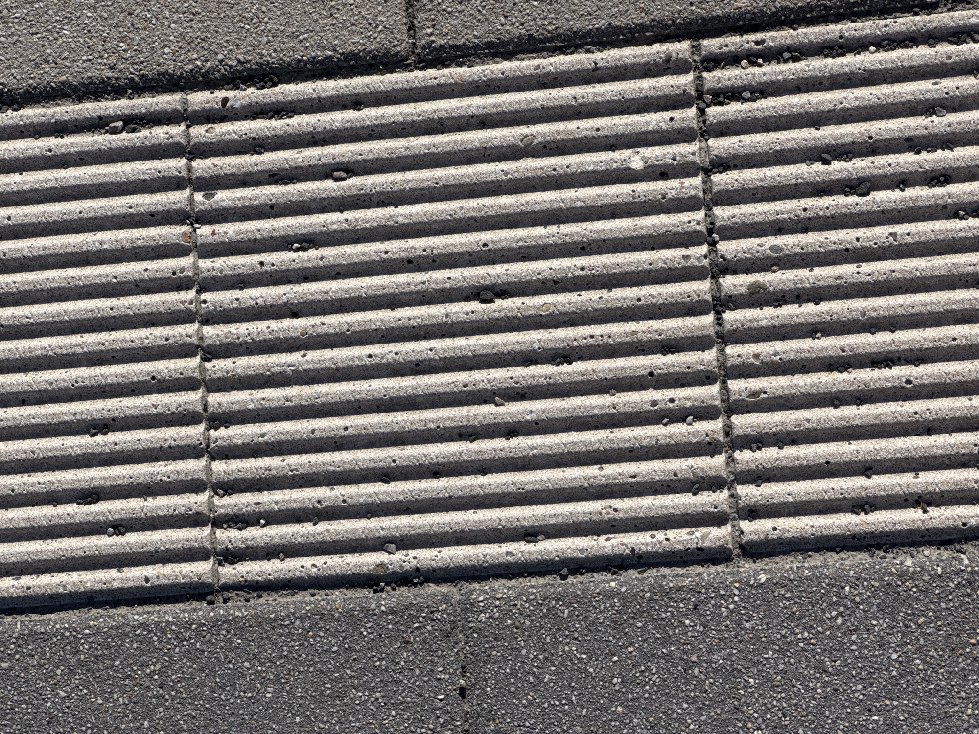 This is a close-up, overhead, forensic photograph of a tactile paving surface adjacent to asphalt paving. The tactile paving consists of light-colored concrete with parallel raised ribs, spaced approximately 2.5 centimeters apart. The ribs run vertically within the frame of the image and are illuminated from the right, casting shadows that emphasize their texture and depth. The concrete surface shows minor imperfections and embedded dark aggregate. To the left of the tactile paving is a dark gray asphalt surface, also with visible aggregate. The boundary between the two surfaces is a clean, straight line. The scene is outdoors, under direct sunlight, casting sharp shadows. This type of tactile paving is commonly found at pedestrian crossings or other areas where visual cues are important for navigation, suggesting a public urban or suburban setting. Given the context provided, this image is from Höhenkirchen-Siegertsbrunn, Germany.