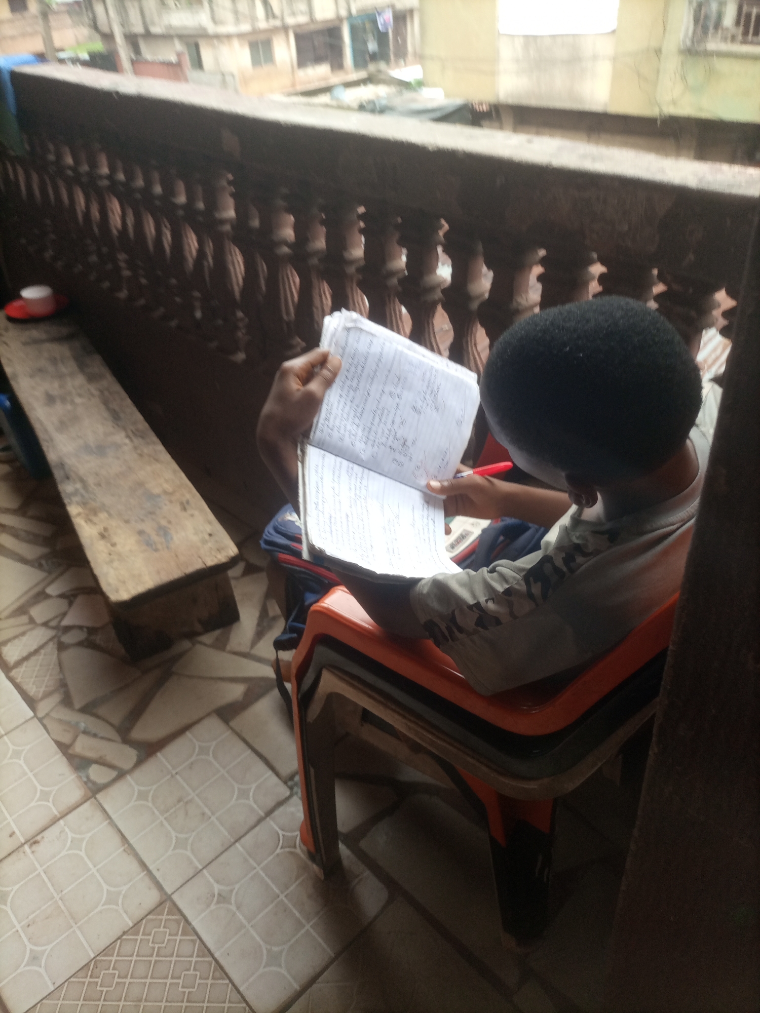 A young Black individual with an afro is seated on an orange plastic chair, angled to the right and facing away from the viewer, engrossed in writing in an open notebook. The individual is wearing a light-colored shirt with a camouflage pattern on the sleeves. In their right hand, they hold a red pen, poised over the notebook. The notebook is open to pages filled with handwritten text. The scene takes place on a covered balcony or porch, with a wooden railing in the foreground. To the left, a wooden bench is visible, and on it, a small white cup on a red saucer. The floor is tiled with a geometric pattern. In the background, blurred buildings suggest an urban environment.