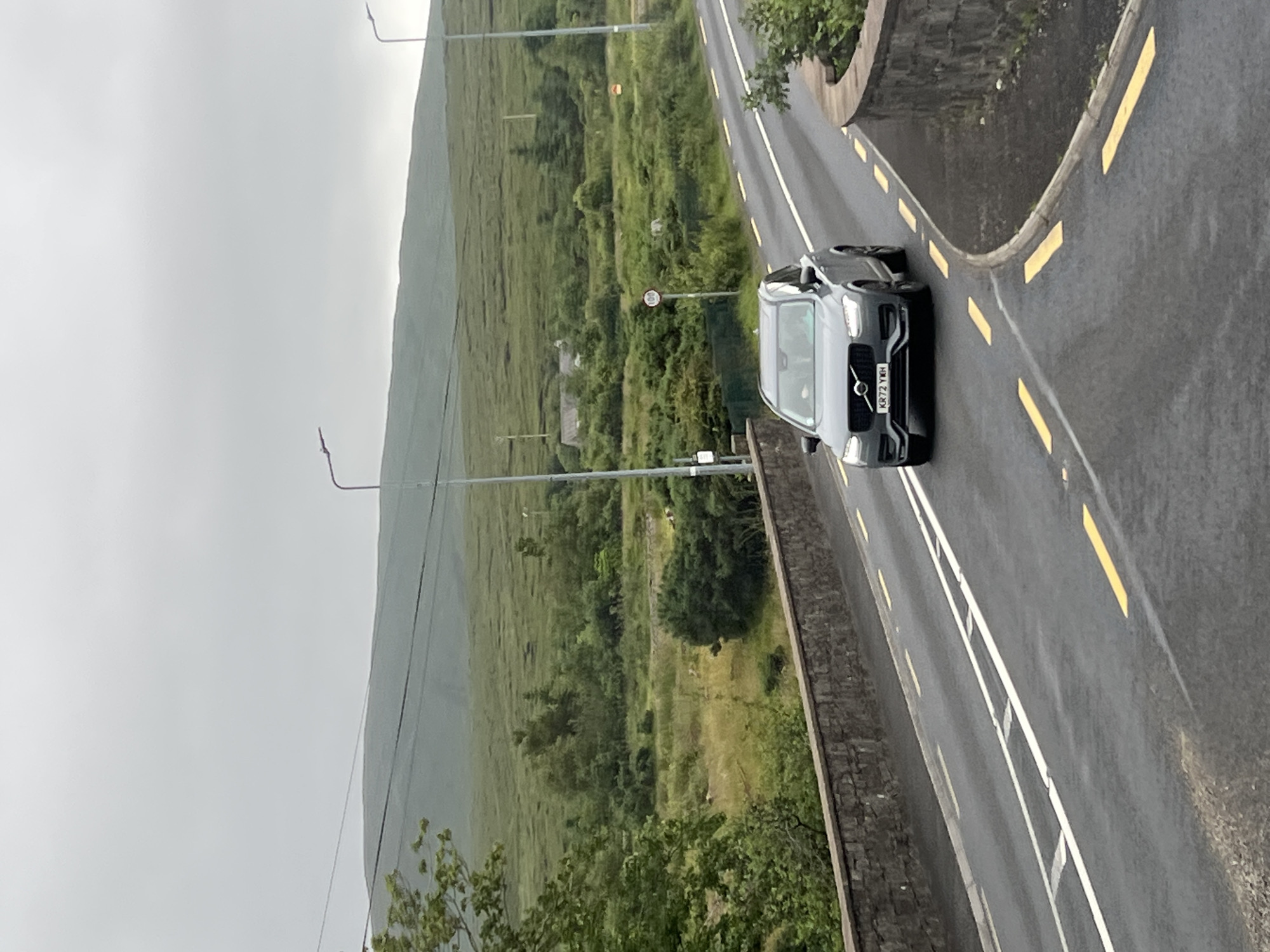 The image shows a dark gray Volvo SUV driving on a wet asphalt road, likely in Gweedore, Ireland. The car is positioned on the right side of the road, with its front facing towards the viewer. The license plate is visible and reads "KR72 YXH". The road has yellow and white markings.
The scene is a rural landscape with rolling green hills covered in lush vegetation. The sky is overcast and appears to be a light gray, suggesting it is either early morning or a cloudy day. The road is bordered by a stone wall on the right, and beyond that, there is more greenery and trees. On the left side of the road, the terrain slopes upwards, covered in dense green grass and some scattered trees, extending to a misty mountain in the background.
A speed limit sign showing "100" is visible on the side of the road in the middle ground, indicating a 100 km/h speed limit. There are also lampposts on the side of the road. The overall mood of the image is calm and perhaps a bit somber due to the overcast weather. The wet road surface suggests recent rain or ongoing light precipitation.