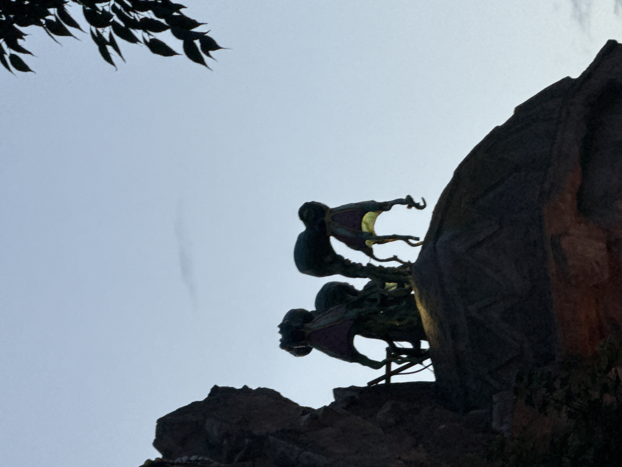 The image depicts a silhouette of figures atop a rocky structure against a pale sky. The figures appear to be stylized, possibly statues or puppets, with multiple limbs and flowing, cape-like elements. Their forms are dark against the lighter background. To the upper right, a portion of dark green foliage is visible, also in silhouette. The overall scene suggests an outdoor setting, likely during twilight or early morning. The image does not contain sufficient information to ascertain a specific location in Vienna, Austria.