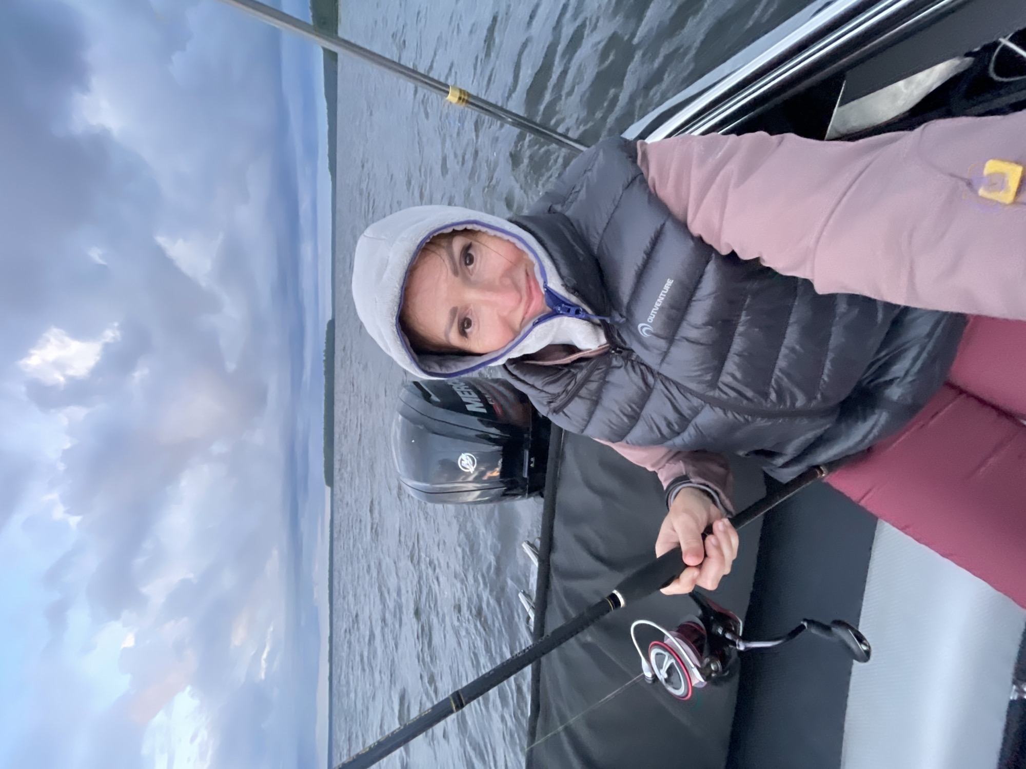 A woman is in a boat on a body of water. She is wearing a gray hooded jacket and a black vest. She is holding a fishing rod in her right hand. The boat has a black outboard motor. The sky is cloudy. The location is Tsvetkovo, Russia.
