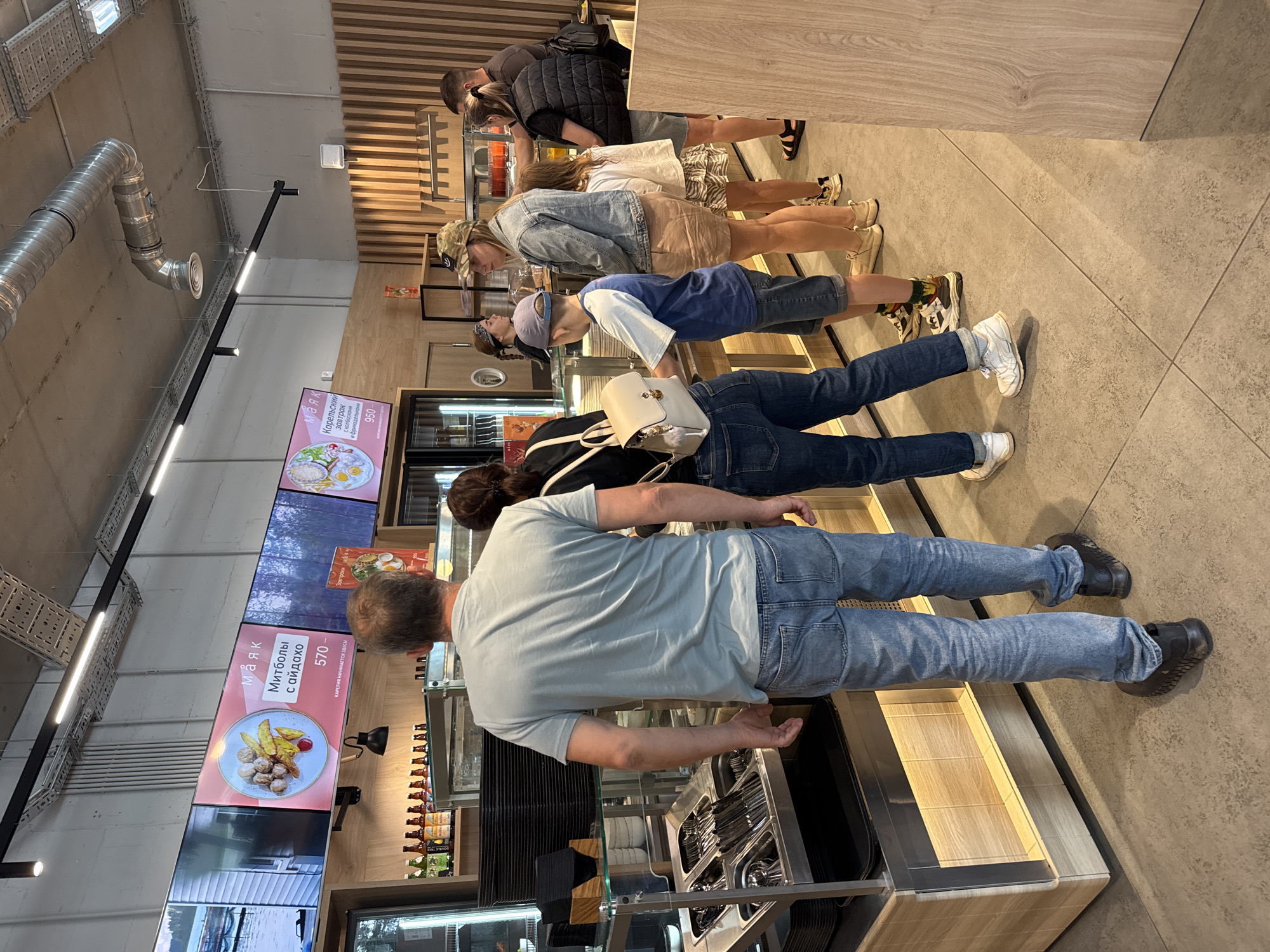 In a fast-casual restaurant setting, likely in Lakhdenpokhya, Russia, a diverse group of individuals are in line to order food. The scene is characterized by a bright, modern interior with wooden accents and digital menu boards displaying food items and prices.
A man in a light blue t-shirt and jeans stands at the front of the line, facing away from the camera. To his right, a woman in a black shirt and jeans is also in line, partially obscured. Further down the line, a young boy in a blue and white t-shirt and shorts, wearing a baseball cap, looks towards the counter. A woman in a denim jacket and beige skirt stands behind him, holding her hand near her face. Another woman with long dark hair in a white t-shirt and a patterned skirt is visible behind the boy. A man wearing a black puffer vest over a t-shirt is standing at the end of the visible line.
The counter area features various food service equipment, including trays, cutlery dispensers, and what appear to be serving dishes. Digital screens mounted on the wall display enticing food imagery, such as meatballs with fries and a breakfast dish. Shelving behind the counter holds bottles of beverages. The overall atmosphere suggests a busy, contemporary eatery.