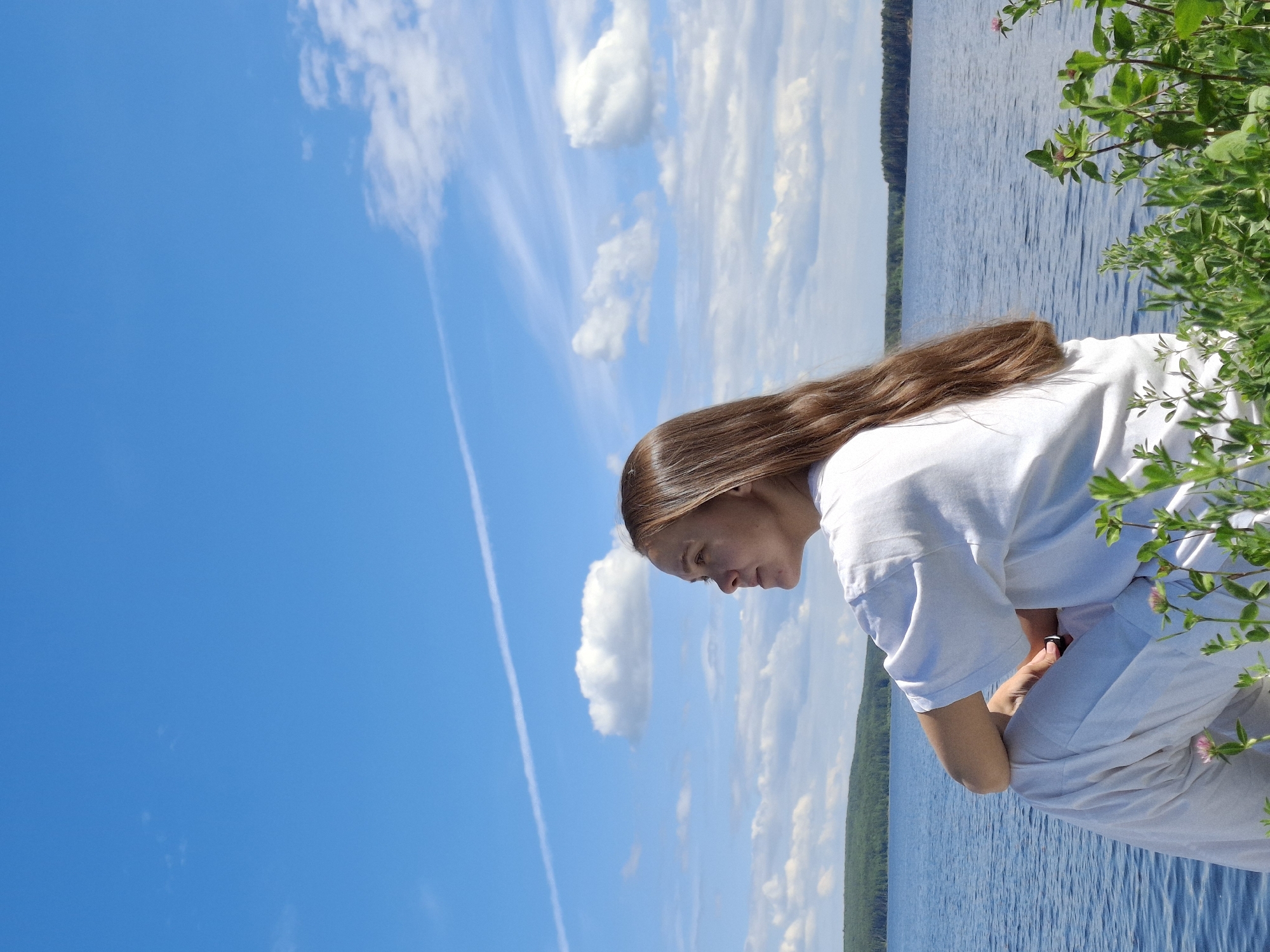 The image displays an adult female seated by a large body of water in the city of Chaikovsky, Russia. The individual, with long brown hair, is dressed in a white t-shirt and white pants, and is positioned in profile, looking left towards the horizon. A small dark object is visible in her right hand, which rests on her knee. The scene encompasses a wide expanse of blue water with minor surface ripples, bordered in the distance by a continuous line of dense green forest. The sky above is a bright blue, featuring scattered white cumulus clouds and a distinct, long white contrail diagonally traversing the upper left quadrant. The immediate foreground includes green plants with small pink blossoms. The environment suggests a clear, sunny day.