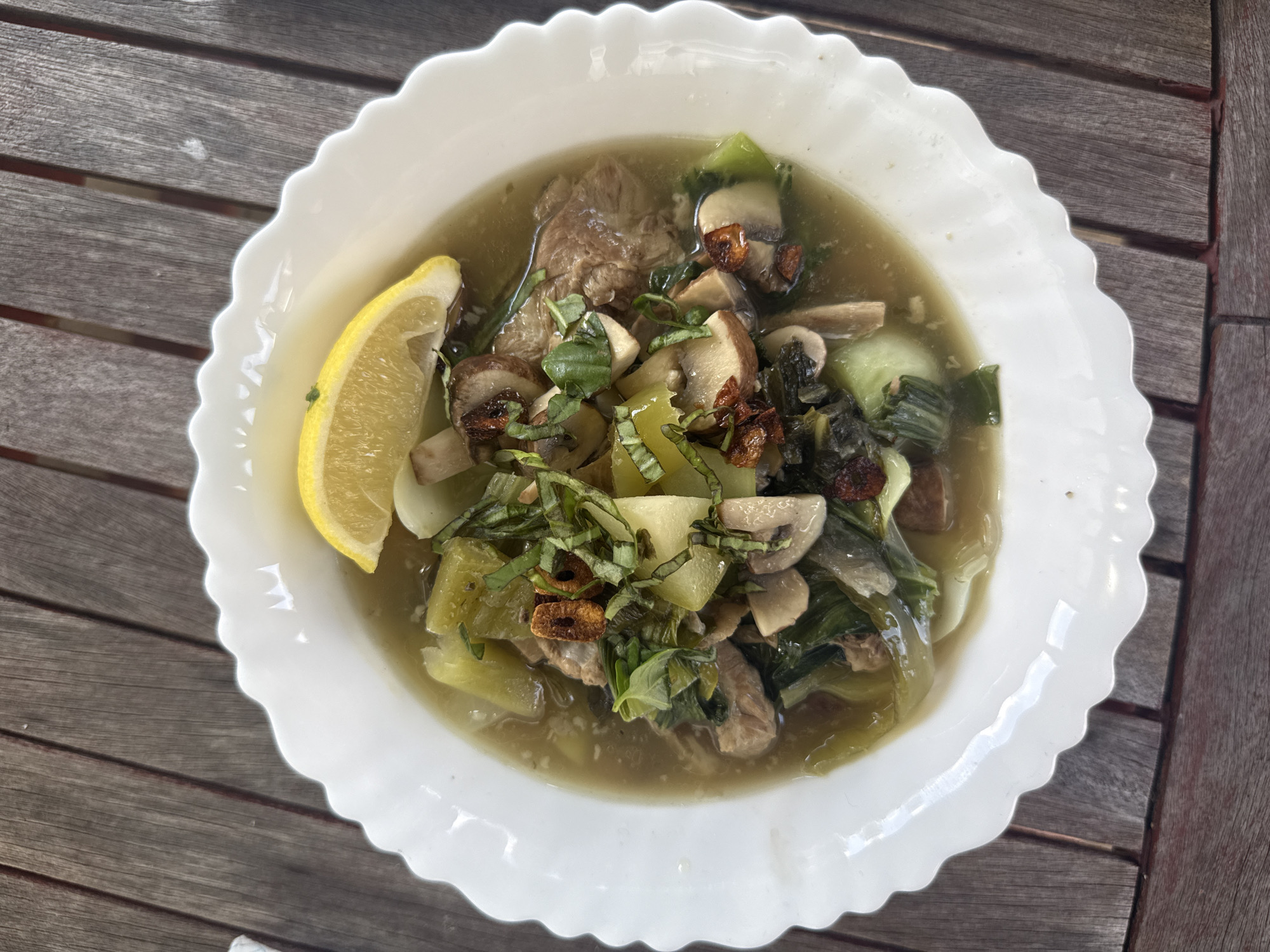 An overhead, close-up shot shows a white, scalloped bowl filled with a steaming broth-based dish. The dish contains chunks of meat, sliced mushrooms, chopped green vegetables that appear to be bok choy, and pieces of potato. The broth is a pale yellow-green color, and the dish is garnished with chopped fresh herbs and crispy fried garlic. A wedge of lemon is perched on the edge of the bowl. The bowl rests on a weathered, wooden outdoor table, suggesting an al fresco dining setting. The lighting indicates it is daytime. There is no discernible indication of the city of London, United Kingdom in the image.