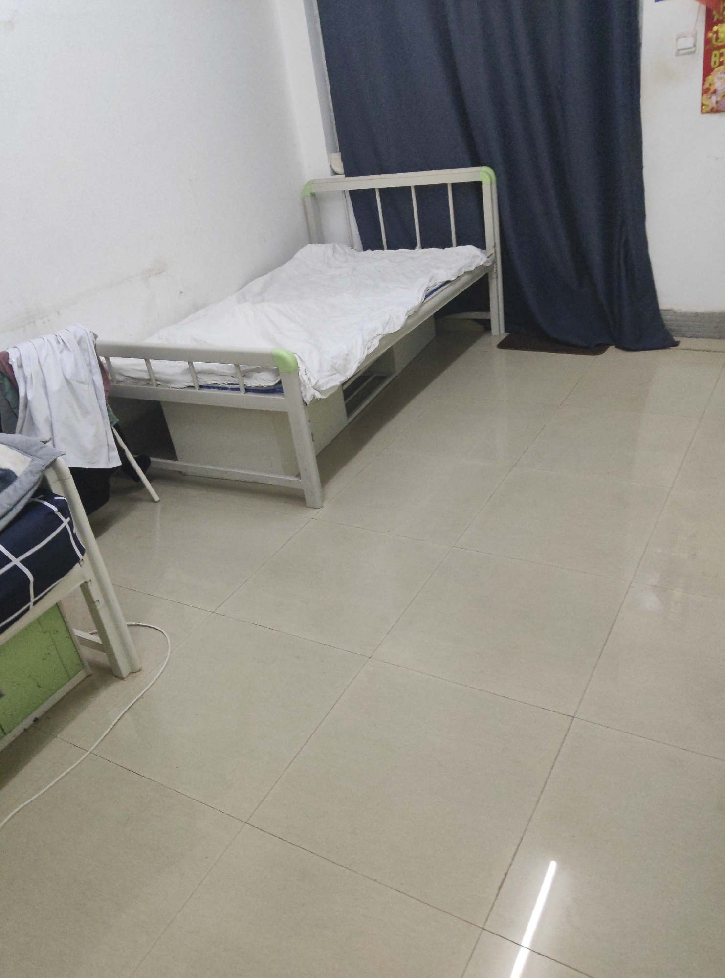 The image depicts a small, sparsely furnished room, likely a dormitory or a basic living space, located in the Chongqing High-tech Zone, China. The room is dominated by two metal-framed beds. The primary bed, closer to the center of the frame, is neatly made with white bedding. Its headboard consists of vertical metal bars. A dark blue curtain hangs to the right of this bed, obscuring a portion of the wall and possibly a window. The walls are painted white, with some faint marks visible, suggesting a well-used space. The floor is tiled with large, square, light beige tiles, reflecting the overhead lighting.
To the left and partially in the foreground, a second bed is visible, lower and with its frame partially obscured by a green panel. This bed has a pillow with a blue and white checkered pattern. Some white clothing is draped over the frame of the first bed, near its foot. A white electrical cord snakes across the tiled floor from the direction of the second bed. The overall impression is one of a functional, utilitarian living environment within a larger residential or institutional complex in the Chongqing High-tech Zone.