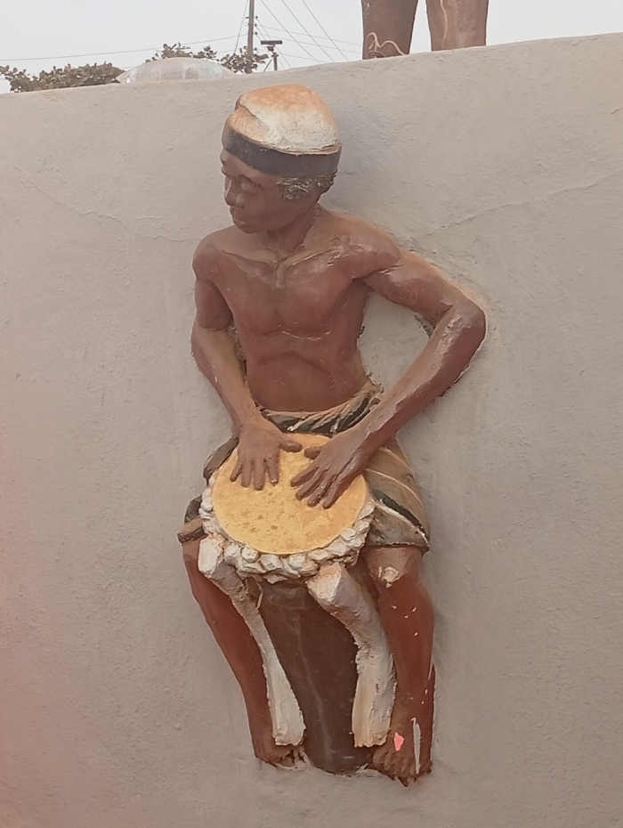A 3D sculpture of a muscular, dark-skinned man playing a round drum. He wears a head wrap and a striped loincloth. His hands are positioned on the drum's surface. The sculpture is mounted on a light-colored wall, seemingly outdoors. Above and partially behind the figure, there are other sculpted forms and a distant view of utility poles and trees. The lighting is soft, creating subtle shadows that highlight the figure's contours.
