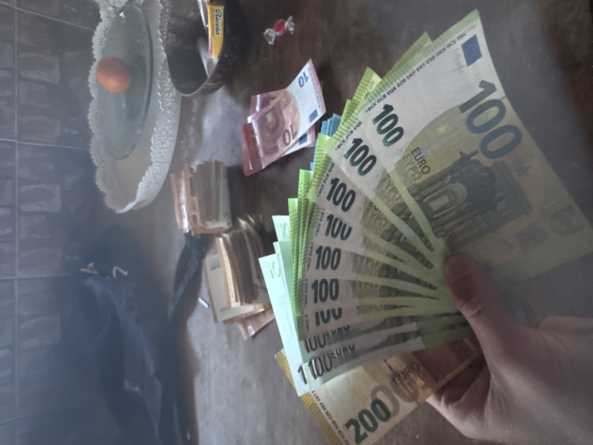 The image shows a person's hand holding a fan of 100 Euro banknotes, interspersed with a few 200 Euro and 10 Euro notes. Stacks of smaller Euro banknotes are also visible on a surface. Other objects in the scene include a glass bowl with an orange, a box of Ricola throat lozenges, and a wrapped candy. A dark object is located to the left of the stack of smaller euro banknotes. The location is unknown.