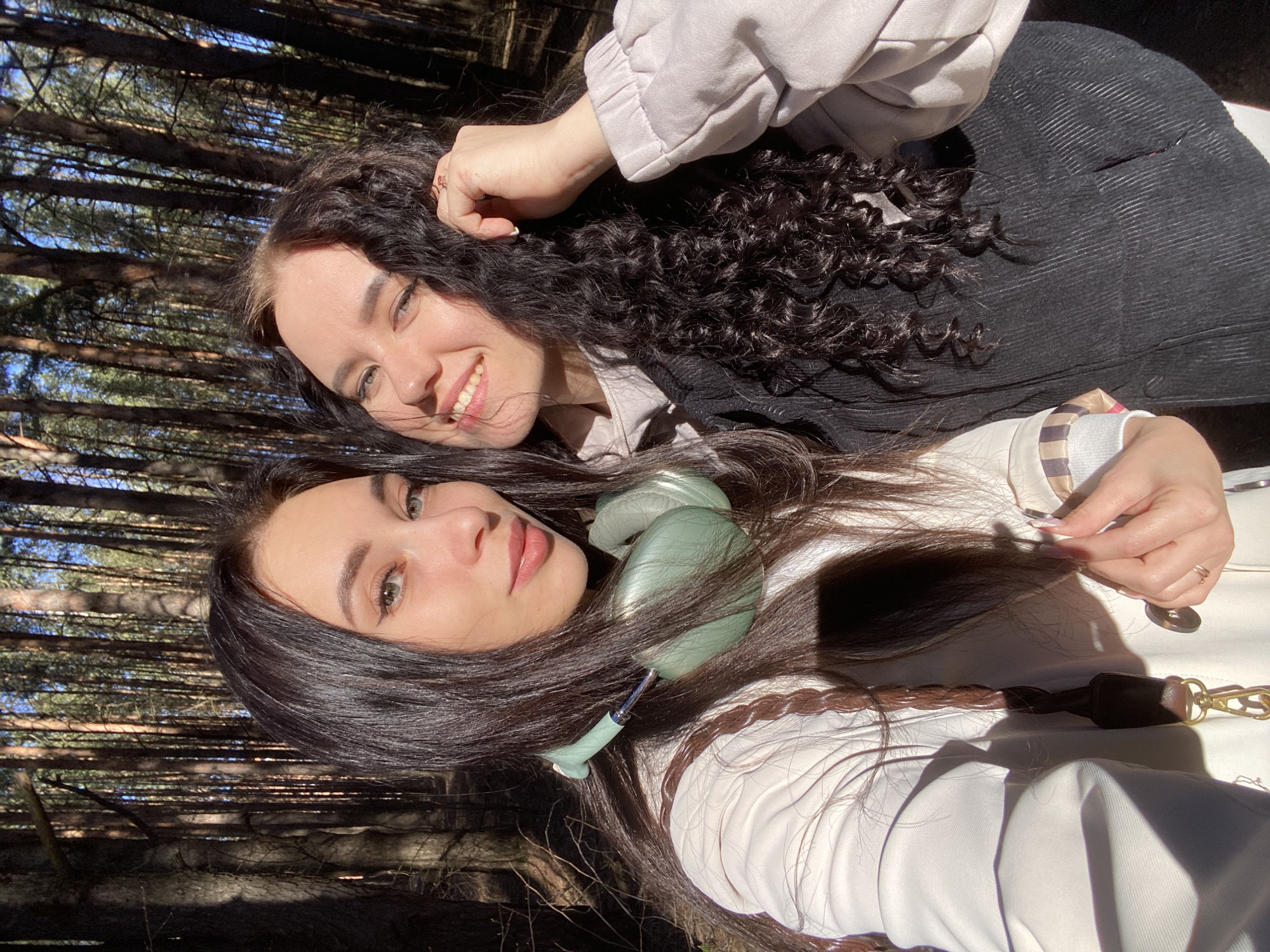 Two women are posing for a photo in a forest setting. The woman in the foreground is wearing light-green headphones and a white coat. The woman behind her has curly dark hair and wears a black top with a light-colored jacket. Trees are visible in the background. The location is Chaikovsky, Russia.