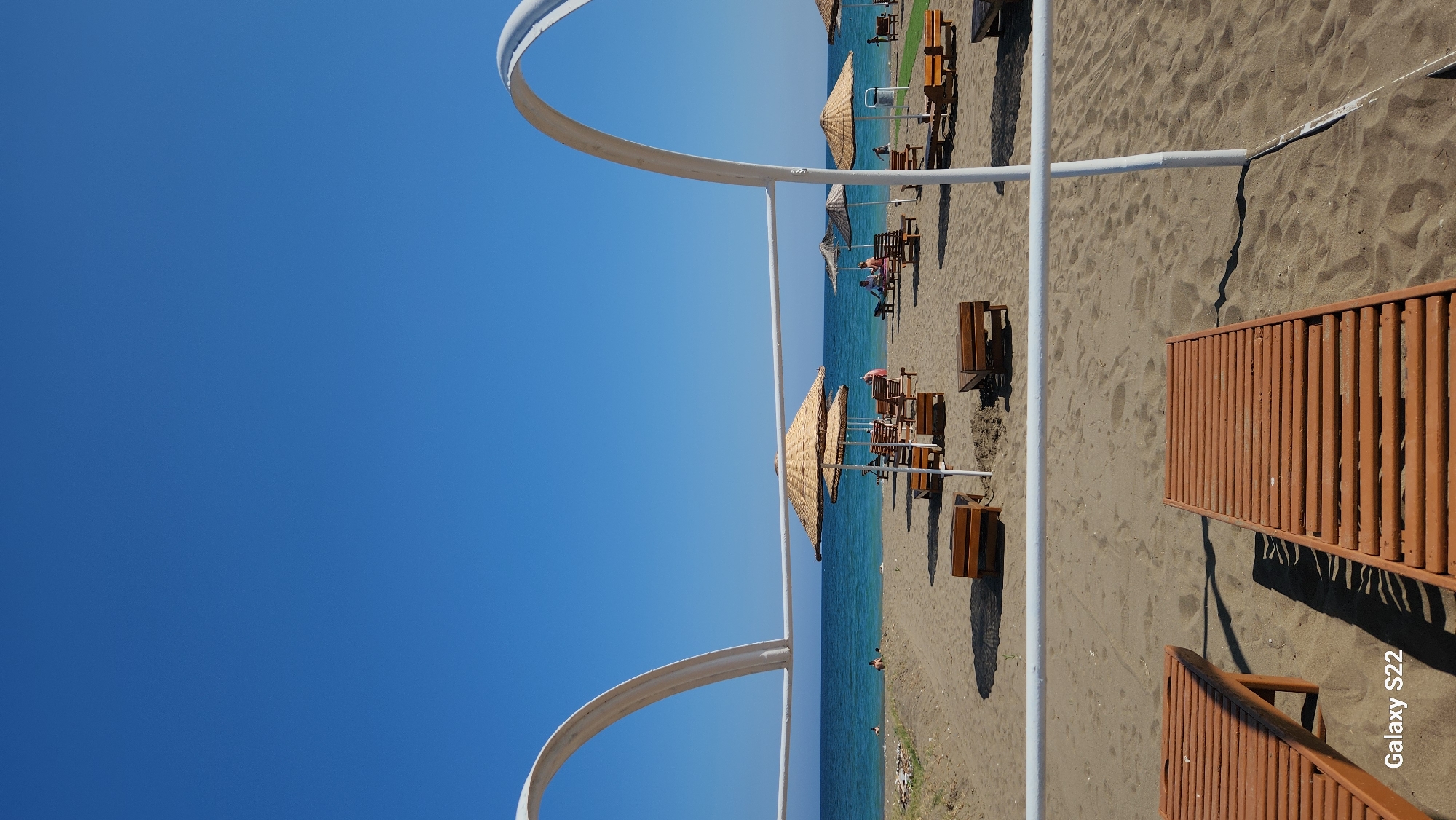 This image depicts a sunny beach scene in Erdemli, Turkey, viewed from a low angle on the sand. The foreground features two partially visible brown wooden sun loungers on textured light-brown sand. A white architectural structure consisting of two curved arches connected by a horizontal bar frames the middle ground. The middle ground presents a sandy beach furnished with numerous straw-thatched beach umbrellas and corresponding wooden sun loungers and small benches arranged in rows. Several individuals are present: two people are in the turquoise-blue sea on the left, while others are seen lounging on sunbeds or sitting on benches on the sand, primarily in the central and right portions of the frame. Some small patches of green grass and a white refuse bin are visible on the far right side of the beach. The background features the calm, deep-blue Mediterranean Sea stretching to the horizon, where a distant landmass is faintly visible on the right. The sky above is clear and intensely blue, devoid of clouds. A "Galaxy S22" watermark is present in the bottom left corner of the image.