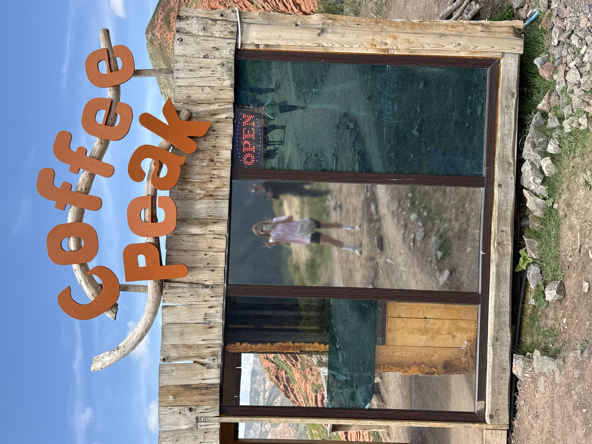 The image shows a sign for "Coffee Peak" made of orange letters attached to a wooden frame, with a glass window behind it. The window reflects the surrounding environment, including a dirt path, people walking, and a mountain in the distance. An "OPEN" sign is visible in the window. The structure is located outdoors, with a rocky and grassy area to the right. Based on the provided context, this scene likely exists in Jeti-Ögüz, Kyrgyzstan.