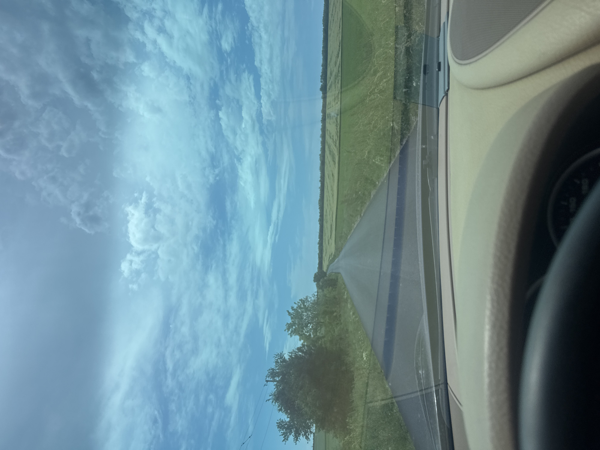 The view from inside a moving vehicle shows a road stretching into the distance through a rural landscape. The sky is light blue with numerous clouds. On either side of the road are fields of green vegetation and some trees. The interior of the vehicle, including part of the dashboard, steering wheel, and door panel, are visible on the right side of the image. The specific location within the city of Glodjevo, Bulgaria, cannot be definitively determined from the visual information available.