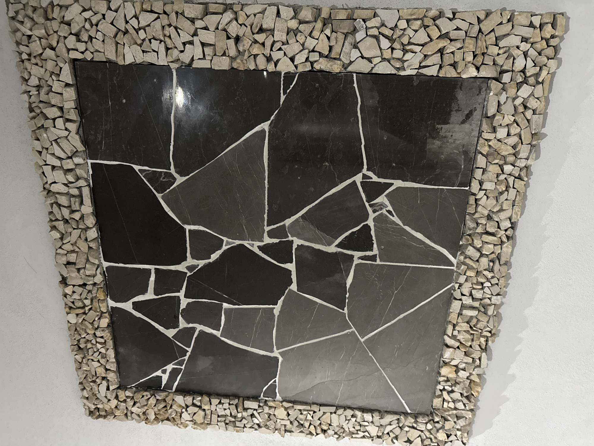 A rectangular decorative panel is mounted on a white wall. The panel features a mosaic design of dark gray stone fragments with white grout lines within a border of small, roughly rectangular, light-colored stones. The setting appears to be indoors. The location is the city of Puebla, Mexico.