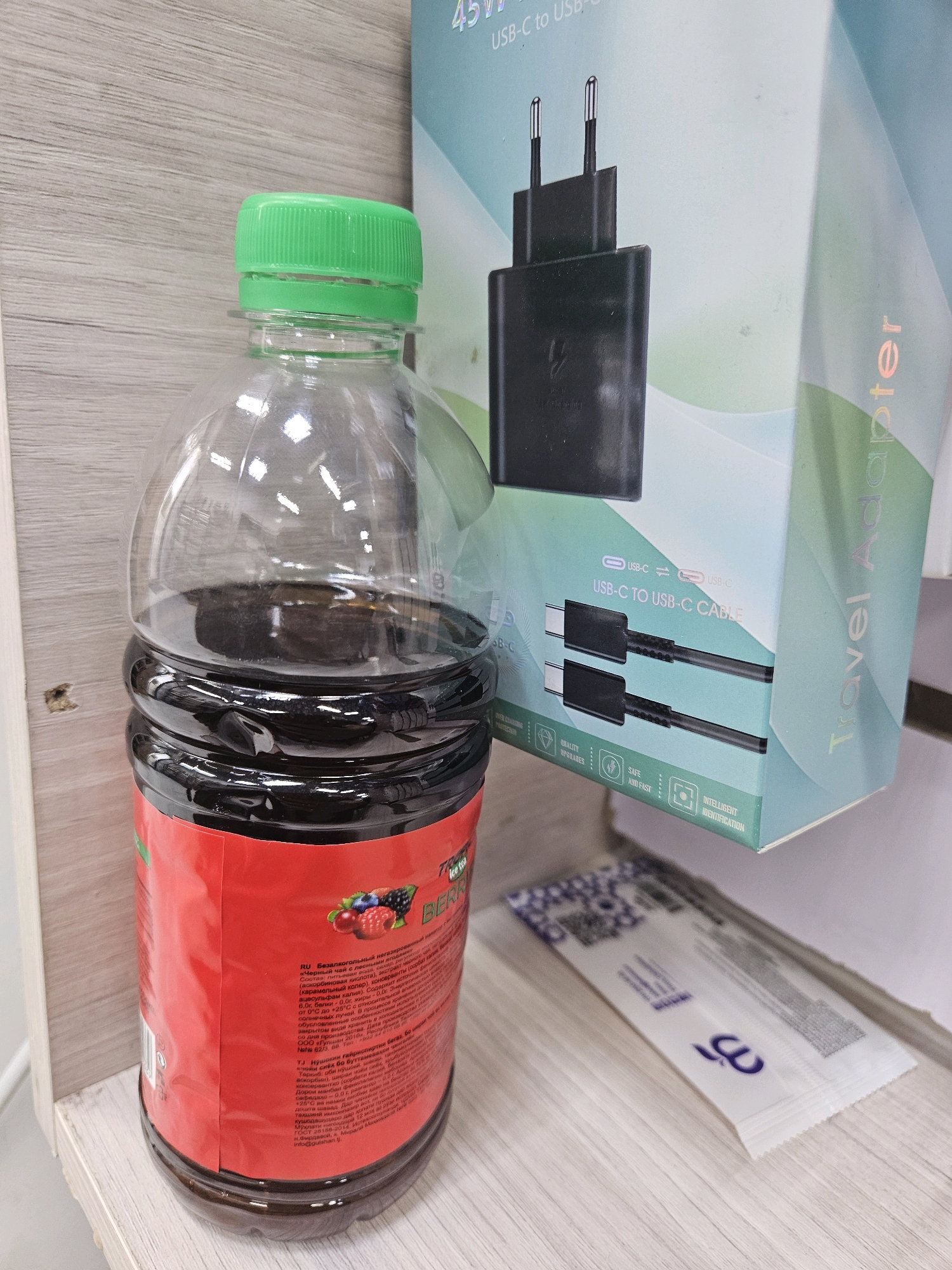 A partially filled plastic bottle of iced tea with a red label is placed on a wooden shelf. Next to the bottle, to the right, is a box for a travel adapter with images of USB-C cables and adapters. A white packet with blue markings is also on the shelf. The background shows a light-colored wooden wall. There are no people in the image. The scene does not provide any information to indicate its location in Dushanbe, Tajikistan.