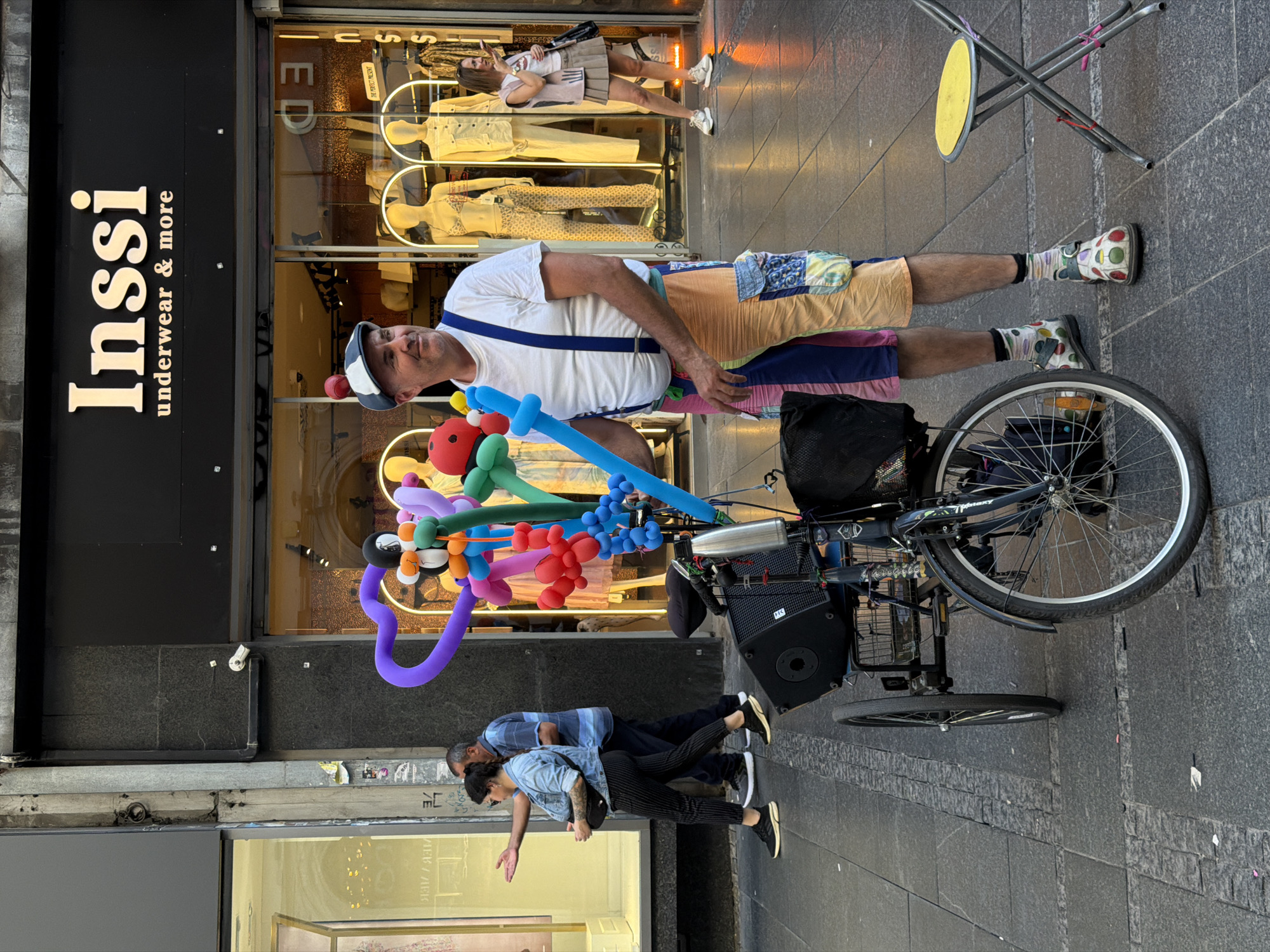 A man stands with his tricycle in front of the "Inssi underwear & more" store on Knez Mihailova street in Belgrade, Serbia. The man is wearing a white T-shirt, blue suspenders, and colorful patchwork shorts. He has a blue and white hat on his head. His tricycle is laden with colorful balloon animals. Two people are walking past him in the lower part of the image, and another person is visible taking a photo inside the clothing store in the background. A yellow folding stool is visible in the upper-right corner. The street and sidewalks are tiled in gray.