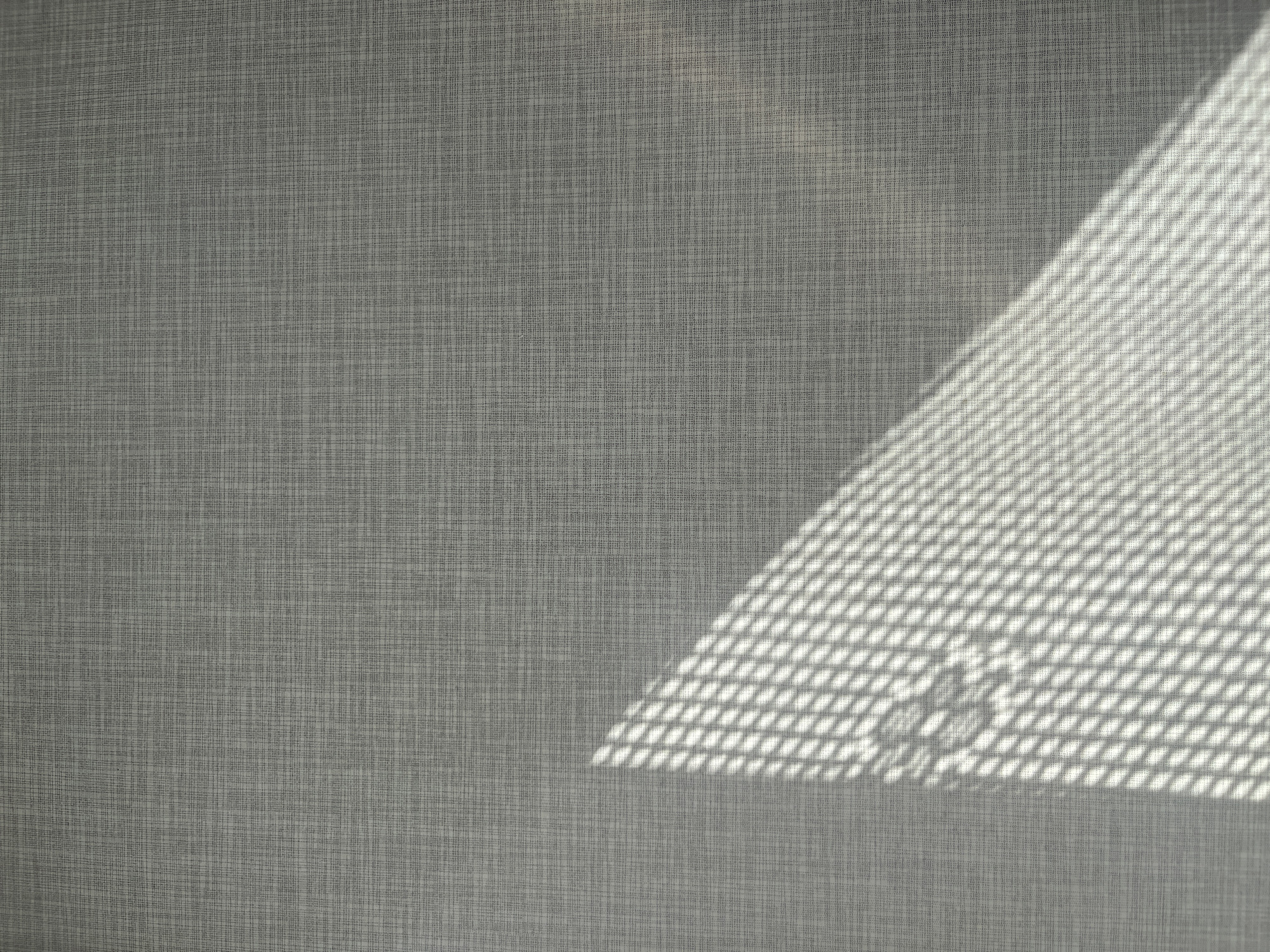 The image shows a close-up of a grey, textured fabric. A shadow falls diagonally across the lower left portion of the fabric, creating a pattern of intersecting lines. The shadow appears to be cast by a blinds or a similar object with a slatted design. The fabric itself has a fine, woven texture. There are no people or discernible objects in the scene, other than the fabric and the shadow. The location in the city of Drohobych, Ukraine cannot be determined from this image.