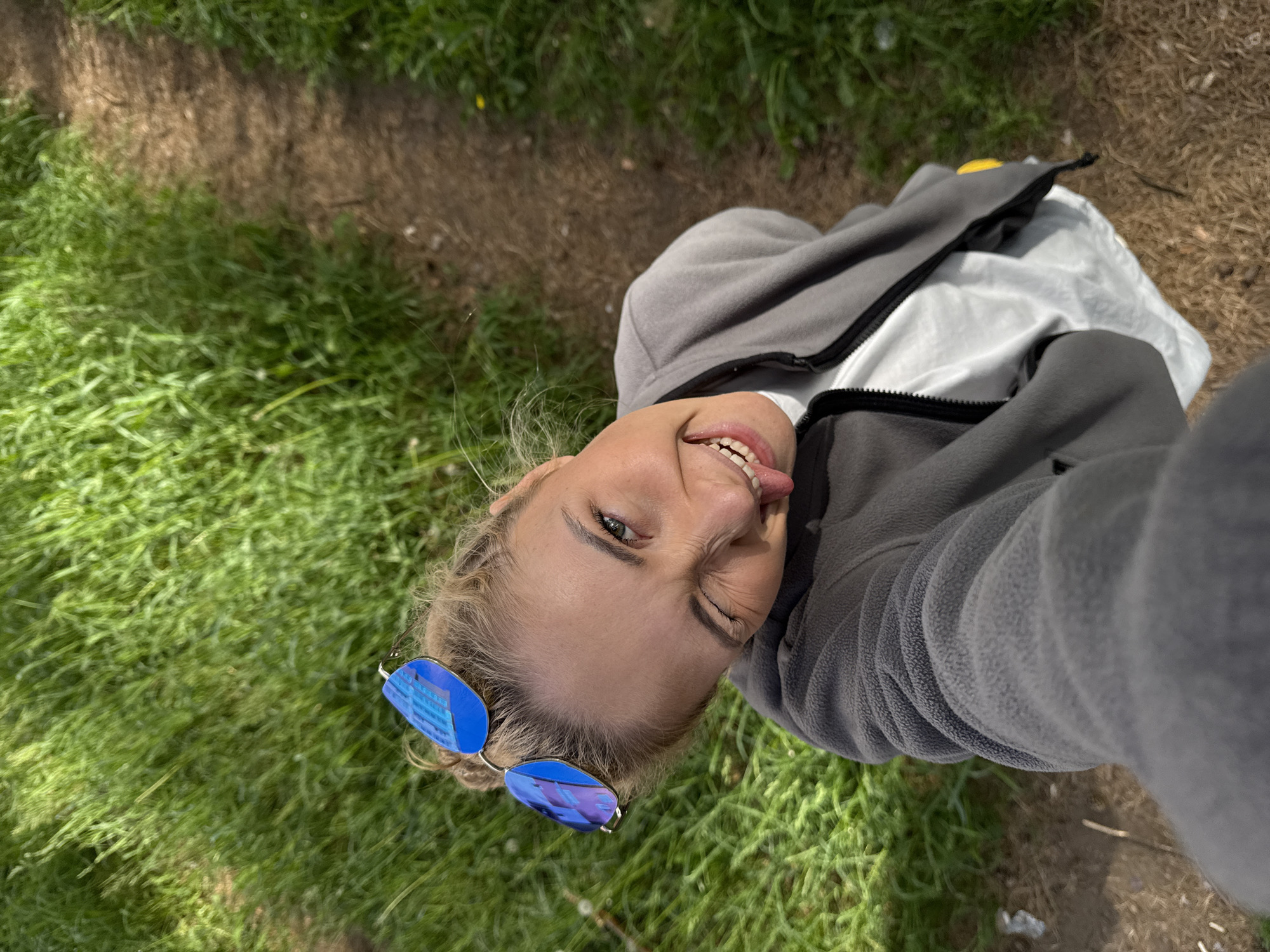 An outdoor selfie captures a young woman from a high-angle, tilted perspective. She has blonde hair and is wearing a grey fleece jacket with a black zipper over a white garment, and blue mirrored sunglasses pushed onto her head, reflecting a blue building-like structure. Her facial expression is playful; she is winking with one eye while sticking her tongue out.
The background is a natural outdoor setting, dominated by vibrant green grass on the left and foreground, contrasting with a dirt path or bare earth visible on the right and in the upper portion of the frame. The scene is brightly lit, indicating a sunny daytime. The overall mood conveyed is cheerful and casual. No discernible text is visible. The provided location context for this scene is Chaikovsky, Russia.