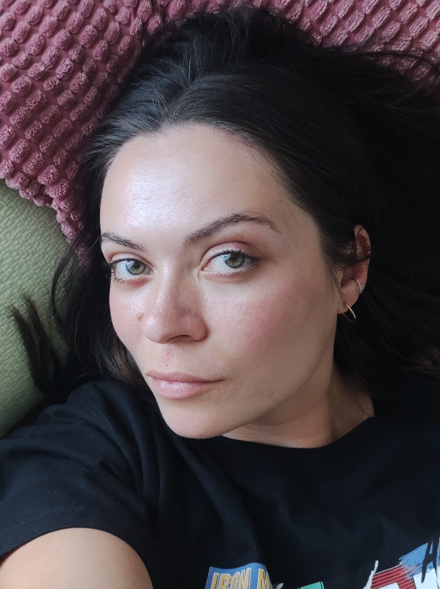 A woman with dark hair and fair skin lies on a light green surface with a textured pink pillow behind her head. She is wearing a black t-shirt and a small gold hoop earring. Her eyes are green, and she has a mole on her cheek. The t-shirt has a graphic design visible at the bottom.
There is no evidence that the image was taken in Chaikovsky, Russia.