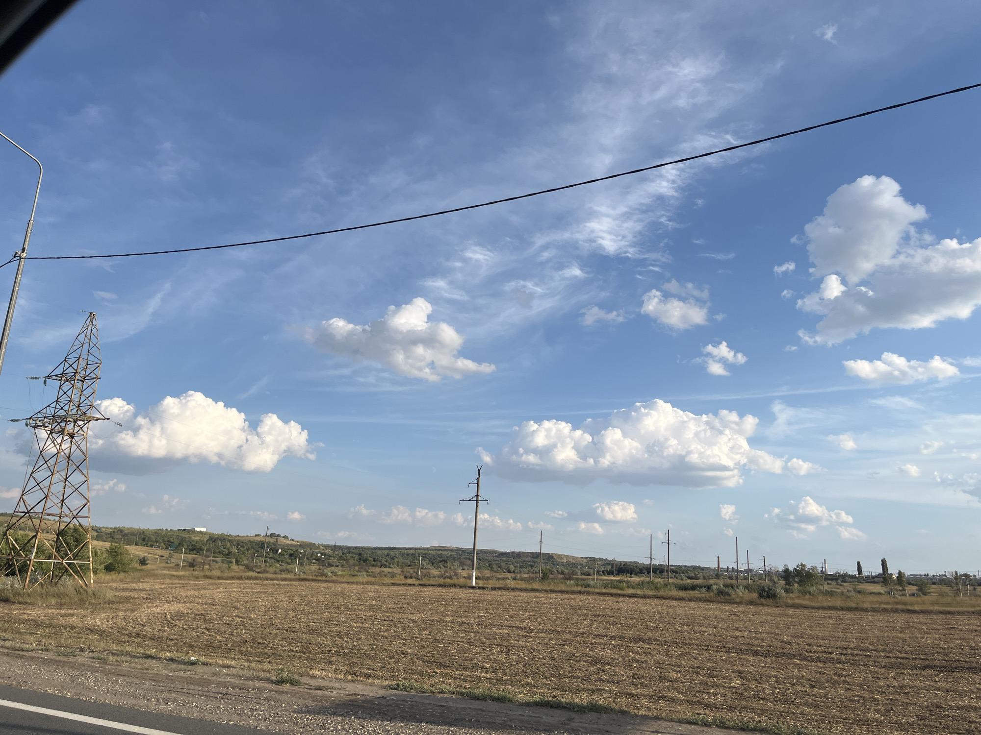 The image captures a rural landscape under a bright blue sky dotted with fluffy white clouds. A large, rusty metal power line tower stands prominently on the left side of the frame, with electrical wires stretching across the sky. In the foreground, a field of dry, harvested crops stretches towards a gently rolling, tree-lined horizon. Several utility poles with electrical wires are visible throughout the middle ground, extending into the distance. The scene appears to be an open field near Ferma, Russia, with evidence of agricultural activity and essential infrastructure.