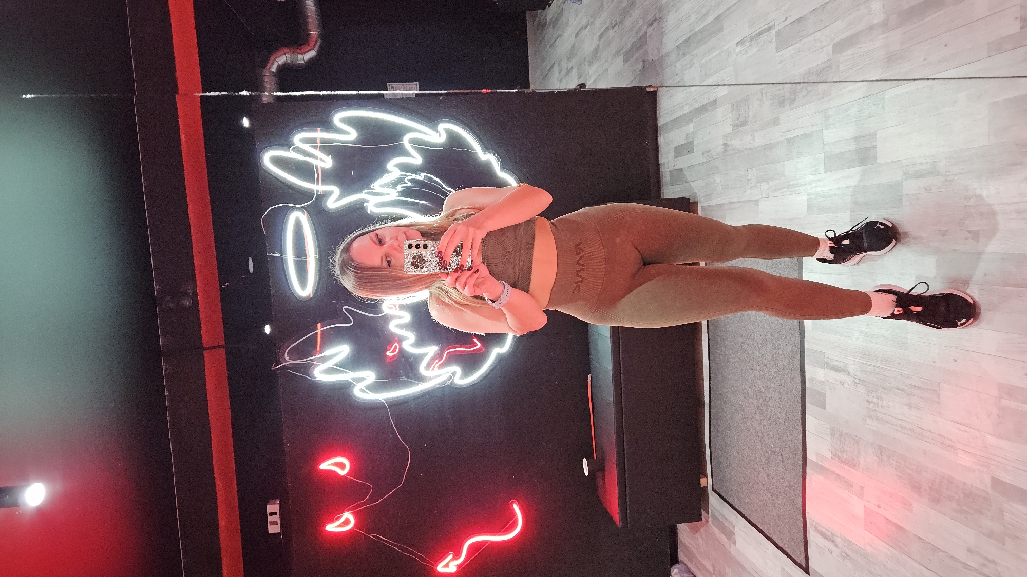 A woman takes a mirror selfie in a gym. The gym has neon lights depicting angel wings and devil horns on the wall. The floor is a light wood-like material and there is a black bench in the background. The woman is wearing a matching olive green workout set and black running shoes. She is holding a phone with a patterned case up to take the photo. There is no indication of location in the city of Sevastopol, Ukraine.