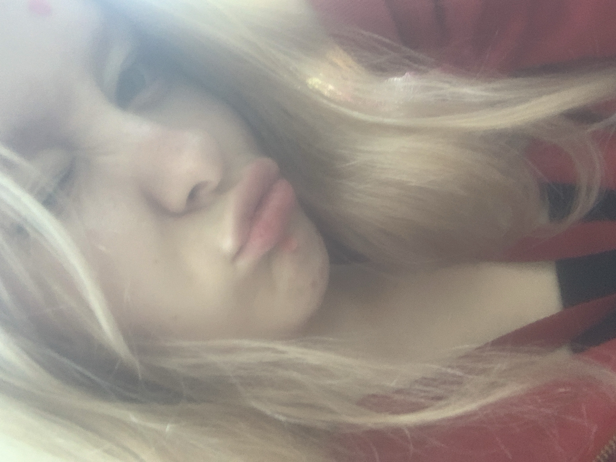A soft-focused, brightly lit close-up image captures a young individual, likely female, with long blonde hair flowing across the frame. The subject's face is shown from a tilted, low-angle perspective, with the prominent features being fair skin, a nose, and distinctly pursed red lips, forming a "duck face" or kissy expression. One eye is partially visible through the light-colored hair and an overall hazy effect.
The individual is wearing a red garment, possibly a jacket or top, visible on the right and bottom portions of the image. The texture of a zipper can be faintly discerned on the lower right edge of the red fabric. The background is indistinct and blurred, offering no specific location cues beyond an implied indoor setting with ample ambient light, suggesting daylight. The image has a dreamy, somewhat overexposed quality with noticeable grain, contributing to its soft aesthetic. No text is visible. The emotion conveyed by the facial expression is playful or expressive.