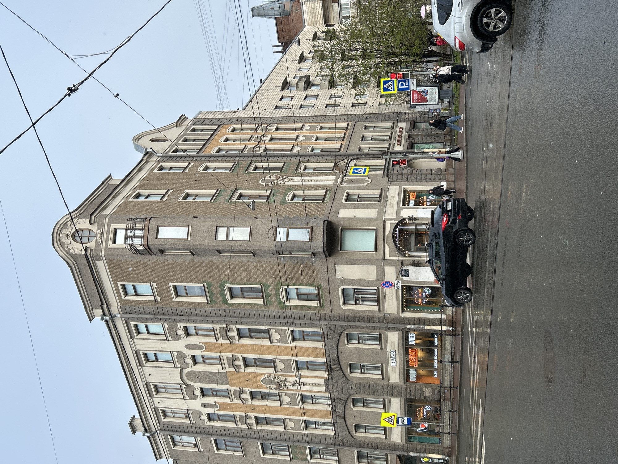 The image captures a street scene in Saint Petersburg, Russia. The architecture is characterized by a tall, multi-story building with a gray facade and ornate window detailing. Street-level businesses, including a bakery with signage in Russian, are visible. A black car is parked along the street, and across the street, pedestrians are using a zebra crossing. Street signs are present, indicating pedestrian crossings and parking availability. Overhead, power lines crisscross the sky. The overall impression is that of a cloudy day in an urban environment. The street is wet, suggesting it has recently rained.