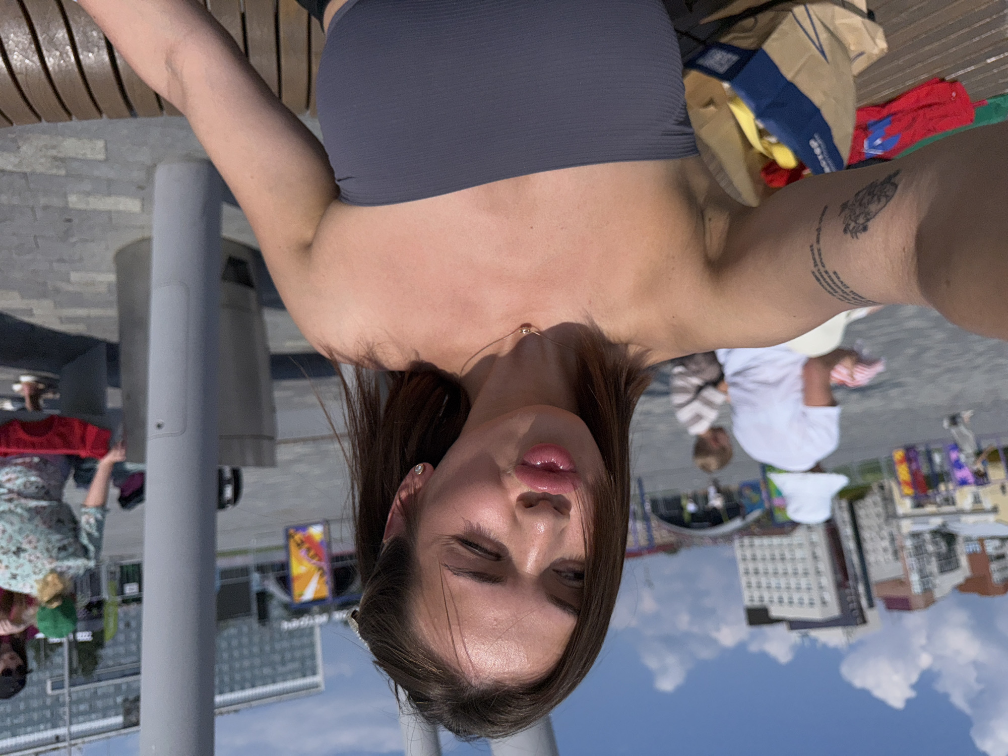 The image shows a young woman with brown hair posing for a selfie outdoors, likely in a public urban space with modern architecture in the background. She is wearing a grey strapless top and a delicate necklace. Her right arm, with a tattoo, is visible, and she has her lips pursed as if blowing a kiss. In the background, several other people are present, some walking and others seated, with what appear to be buildings and a large metal structure dominating the scene. The sky is blue with scattered white clouds, suggesting it is a sunny day. This scene is consistent with a location on the embankment of the Kama River in Perm, Russia, which features modern architectural elements, public spaces, and recreational areas.