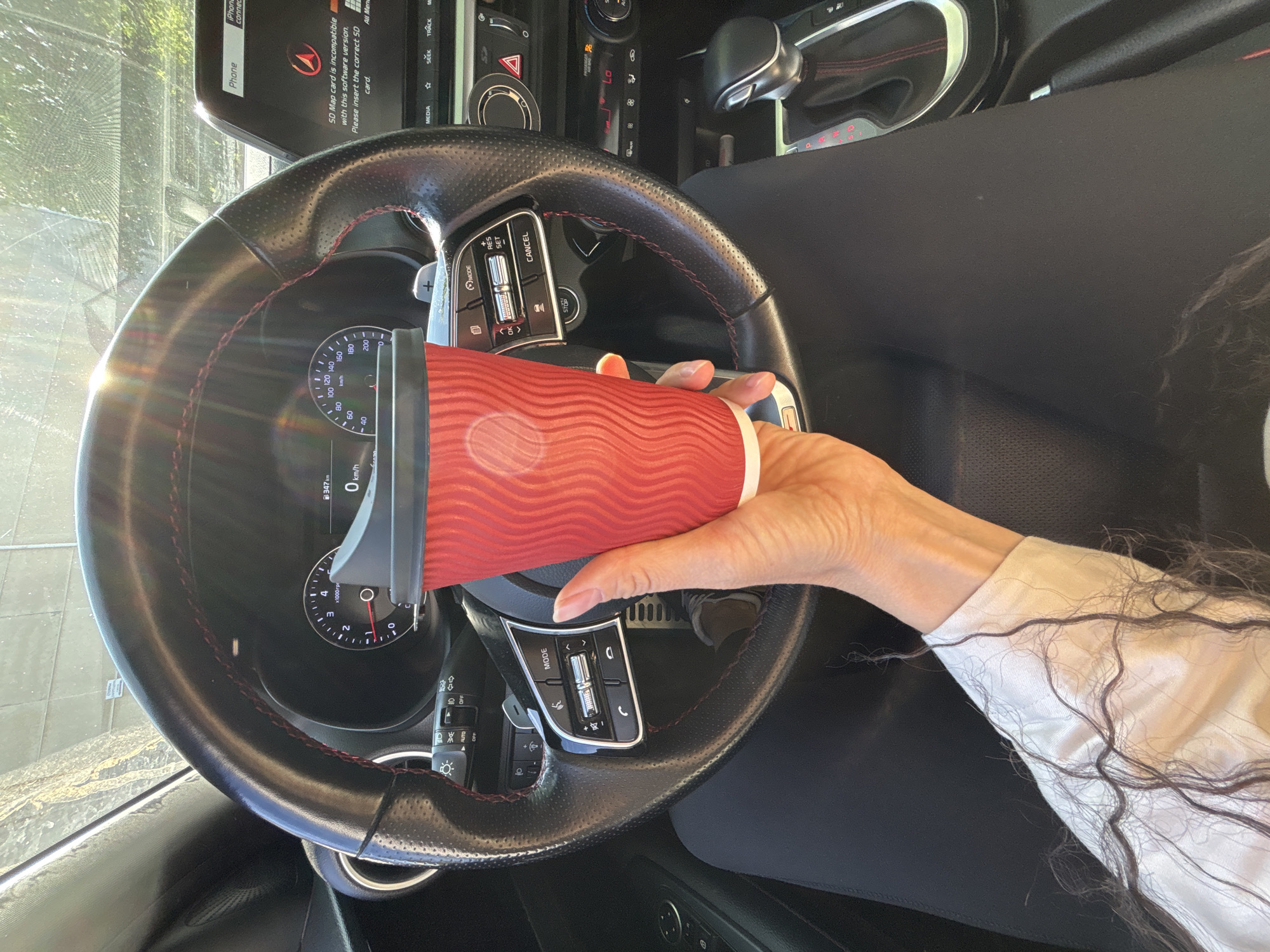 The image provides a first-person view from inside a modern car, likely situated in Chaikovsky, Russia, on a bright, sunny day. A person's right hand and forearm are prominently featured, holding a red disposable coffee cup with a black lid. The person wears a white long-sleeved shirt, and long, dark, wavy hair is visible near their arm.
The car's interior is detailed, focusing on the black leather steering wheel, which features red stitching and perforated grips. Numerous controls are integrated into the steering wheel, including buttons labeled "MODE," "RES," "SET," "CANCEL," alongside phone-related controls for answering/ending calls, adjusting volume, and skipping tracks.
Behind the steering wheel, the dashboard instrument cluster is visible, showing a speedometer indicating "0 km/h" and a digital display reading "347 km," likely an odometer or trip meter. A tachometer marked "x1000rpm" is also present. The infotainment screen in the upper center displays several messages, most notably: "SD Map card is incompatible with this software version. Please insert the correct SD card." Other text on the screen includes "Phone," "iPhone connect...", "All Men," "MEDIA," "SEEK," and "TRACK."
Portions of the center console are also discernible, including a gear shifter with red accents and climate controls set to "LO" and "88." Bright sunlight glares off the windshield and dashboard, reinforcing the daytime setting. The exterior view through the windshield is indistinct but suggests an outdoor environment. The "km/h" unit on the speedometer is consistent with the provided location context of Chaikovsky, Russia.