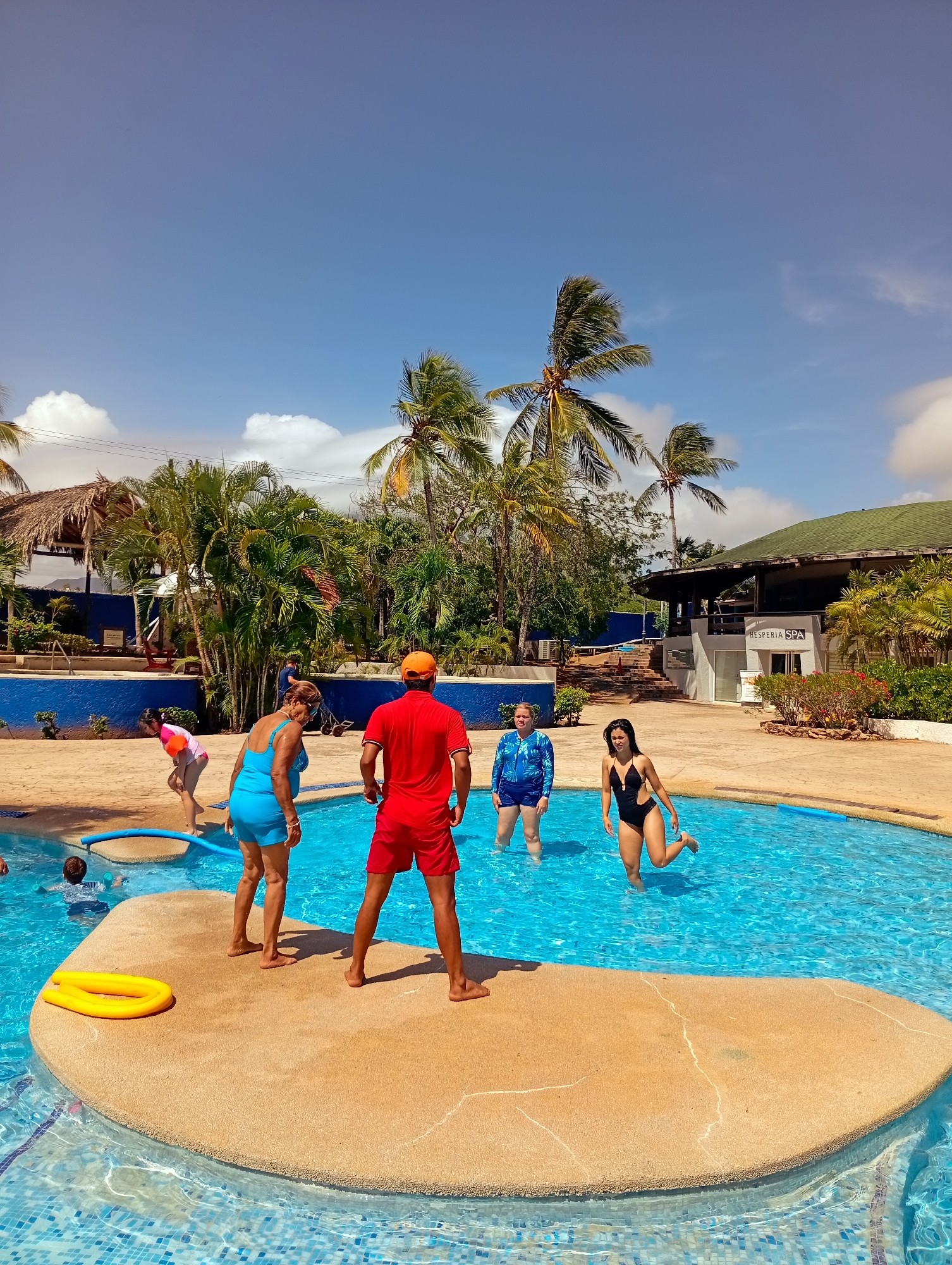 The image depicts a scene at a swimming pool in Playa El Agua, Venezuela. A group of people is gathered around the pool, some standing on a small island within the pool. An adult male wearing red shorts and a red t-shirt and an orange baseball cap stands on the island. Two women, one wearing a blue swimsuit and the other a blue patterned swimsuit, are also on the island, as is a woman in a black swimsuit, and a young child in a pink swimsuit and a boy with his head in the water. A yellow swim noodle sits on the island. The pool is surrounded by palm trees and buildings, one with signage that reads "HESPERIA SPA". The sky is clear and blue with some scattered clouds.