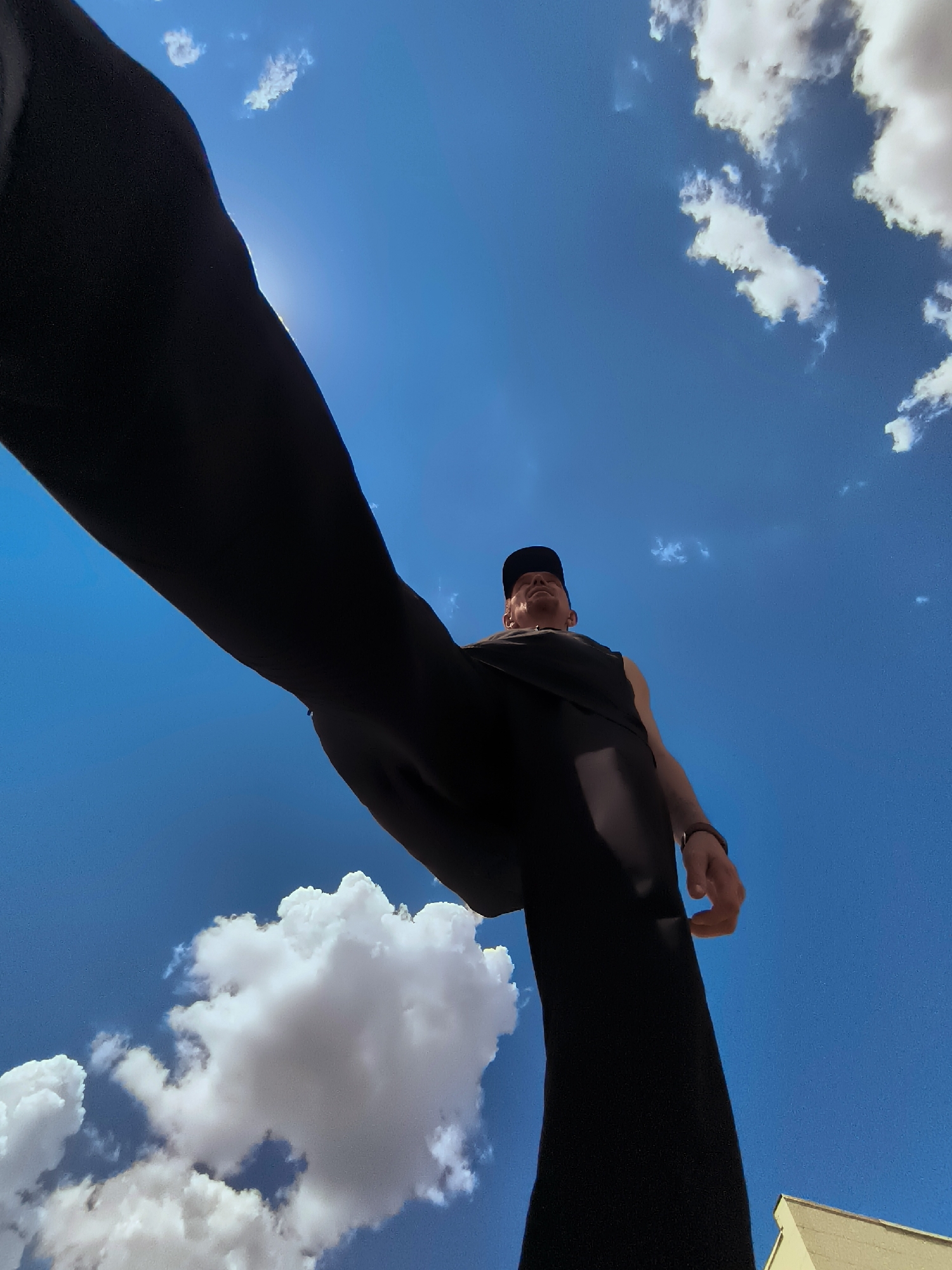 A low-angle, wide shot captures a person standing against a bright blue sky with scattered white clouds. The person, wearing a black sleeveless shirt, a black baseball cap, and dark pants, is seen from the waist down and left arm side. Their head is tilted upwards, and they appear to be looking towards the sky. A thin, dark band is visible on their left wrist. A hint of a light-colored building is visible in the lower right corner. The image's perspective suggests the photo was taken by someone holding a camera at ground level or below the subject. There are no discernible landmarks to identify the specific location in Karaganda, Kazakhstan.