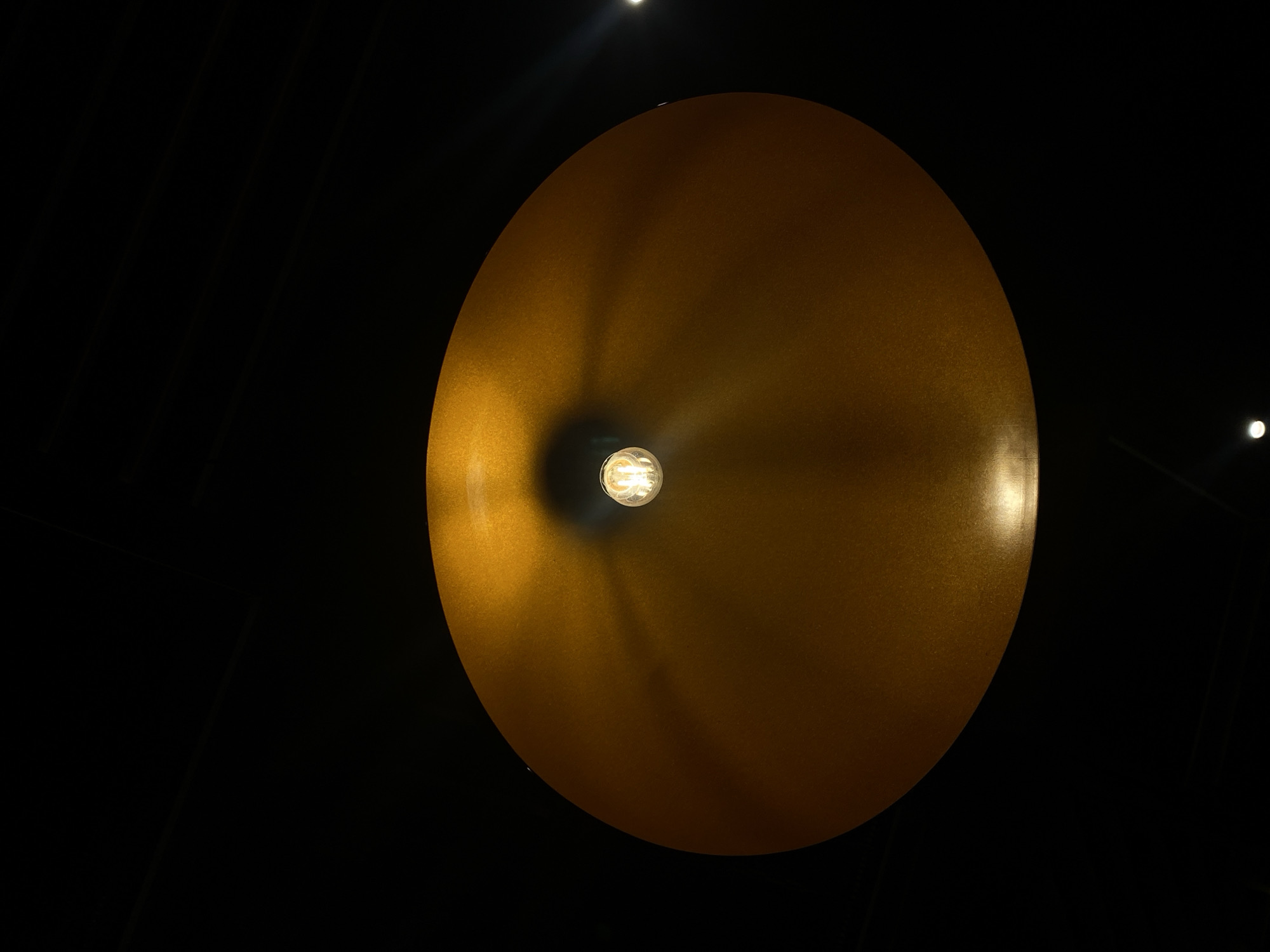 A low-angle shot focuses on a golden, disc-shaped lamp with a visible light bulb in its center, suspended against a dark background. Two small, out-of-focus lights are visible in the background, one near the top left and another on the right. The lamp's surface shows varying light reflections, suggesting a textured or slightly uneven finish. The location is Kazan, Russia.