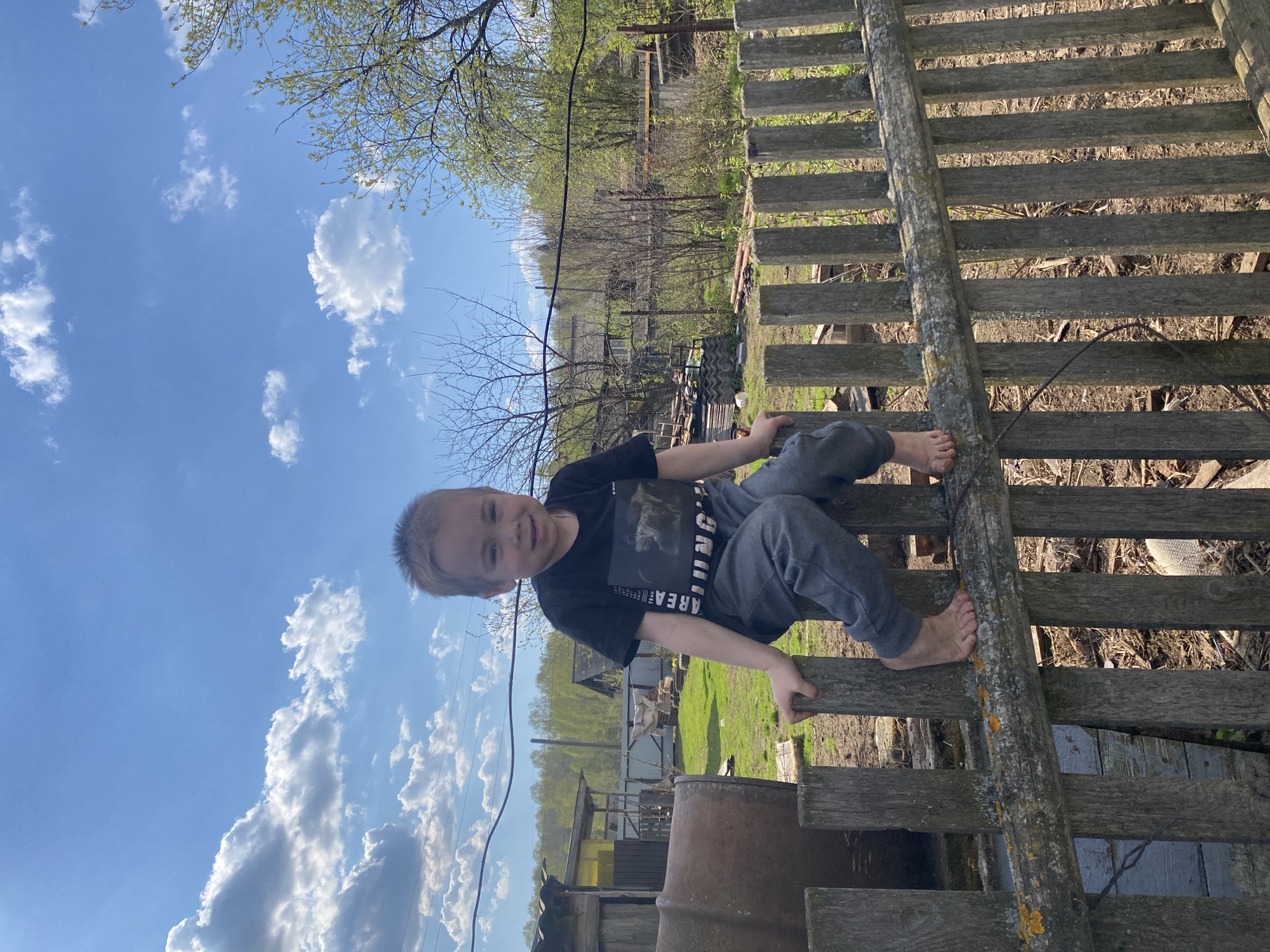 A young boy with short blond hair is perched barefoot on a weathered wooden fence. He wears a dark t-shirt and grey pants. The backdrop consists of a partly cloudy sky and rural scenery, featuring trees, grass, and structures including a metal barrel and a building with a yellow tarp visible. The image suggests an outdoor setting, possibly a yard or countryside area. The location mentioned, Узяр, Russia, cannot be definitively determined from this image alone.