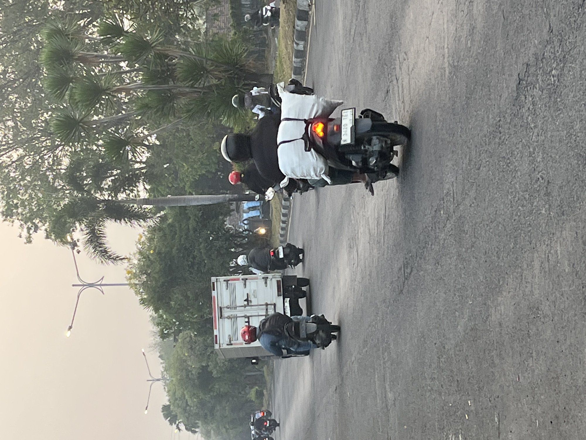 In Kludan, Indonesia, a grey, overcast sky hangs over a two-lane asphalt road. Multiple motorcycles are in motion. In the foreground, a motorcyclist wearing a black jacket and a dark helmet carries a large, white bundle secured to the back of their motorcycle. The motorcycle's license plate reads "N 3420 TEI". To the left and slightly ahead, another motorcyclist with a red helmet is positioned behind a white truck. The truck has a white cargo box with red accents. Further in the distance, other motorcycles and vehicles are visible on the road. Trees, including palm trees, line the sides of the road, with streetlights visible in the background. The overall scene depicts traffic on a roadway.