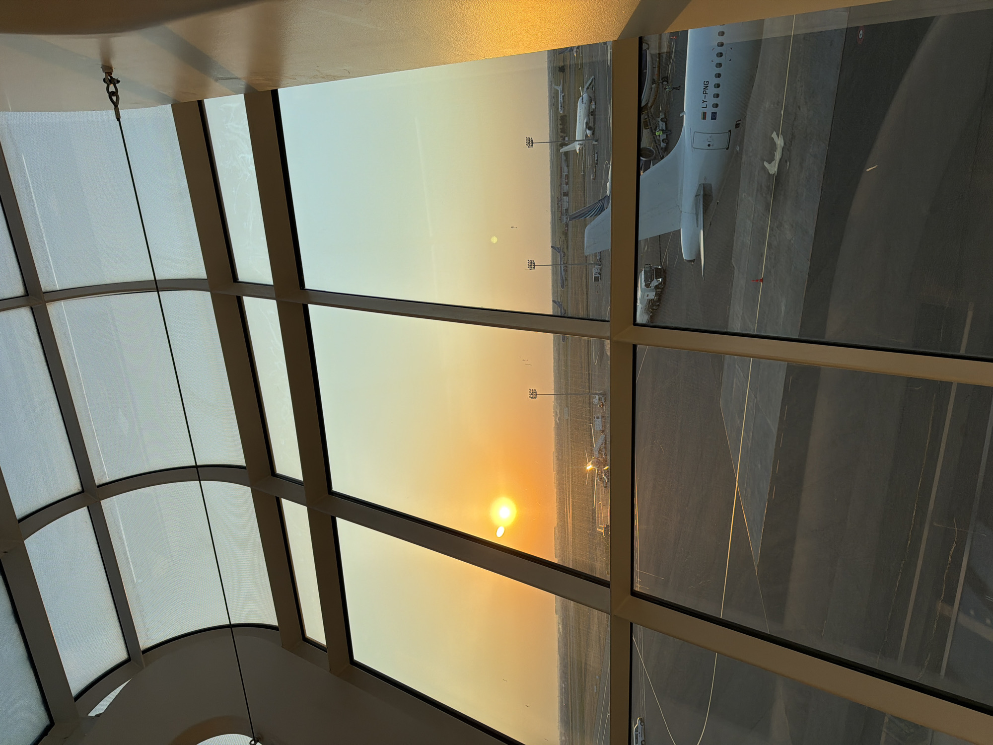 The image captures a view from inside an airport terminal, likely within the city of Khazar Raion, Azerbaijan, during sunset. A large, curved glass ceiling with segmented panels lets in the warm, orange glow of the setting sun. Through the expansive windows, an airport tarmac is visible. Several airplanes are parked, including a white passenger jet in the foreground with the registration "LY-PNG" visible. A fuel truck is positioned near one of the aircraft. The tarmac is marked with various lines and indicators. In the distance, more aircraft and airport infrastructure, such as lighting towers, can be seen. The overall scene depicts the operational activities of an airport at dusk, with the natural beauty of the sunset adding a serene element. There are no discernable people in clear focus.