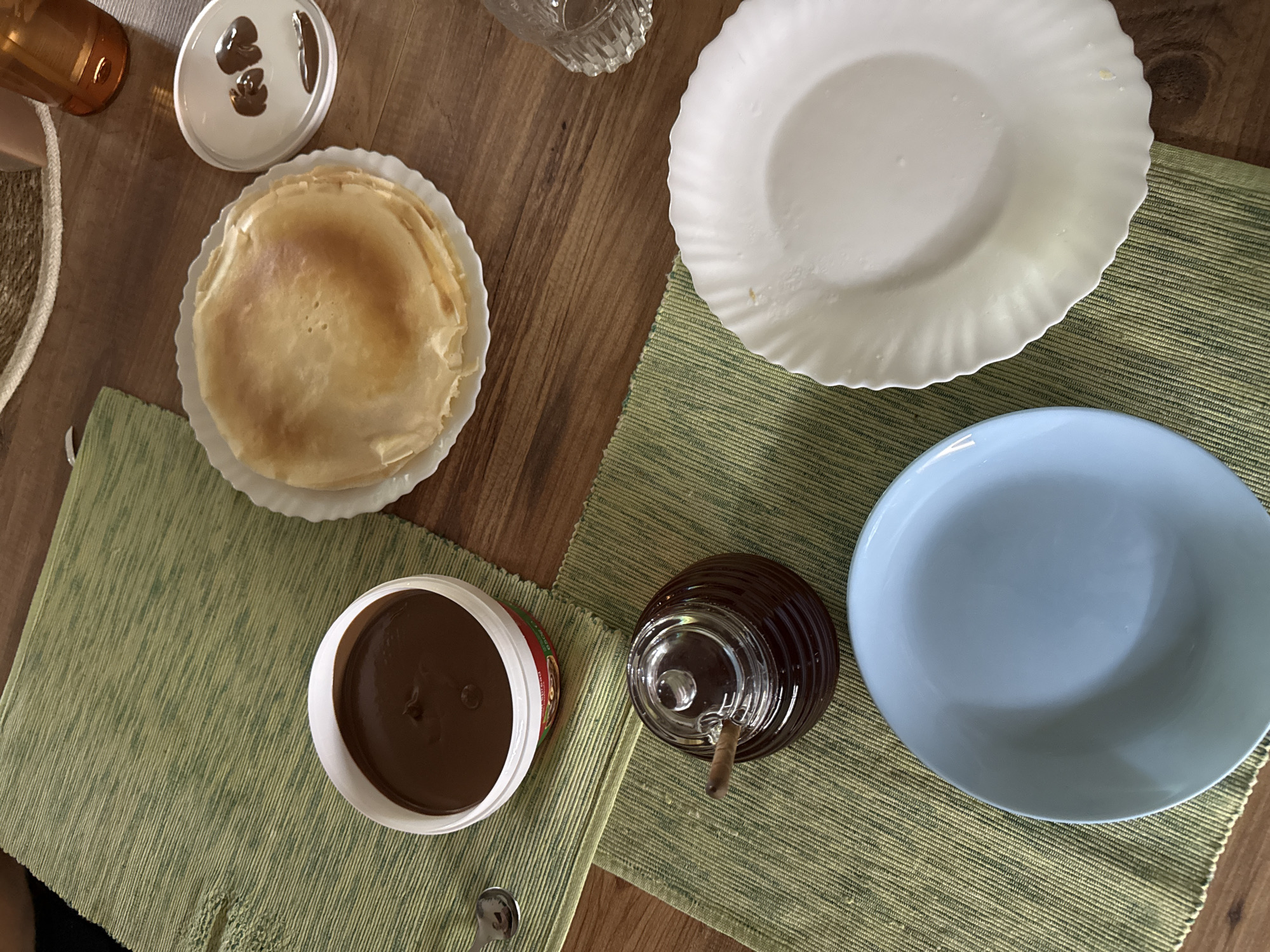 A breakfast scene is set on a wooden table covered with green placemats. In the center of the table, a stack of golden crepes sits on a white paper plate. To the left, a white plastic tub filled with chocolate spread is partially visible, with a small spoon resting on the placemat beside it. To the right of the crepes, a small white lid with dollops of chocolate spread is placed on the table. Further to the right, a glass of water sits on the wooden surface. In the foreground, a light blue plate is positioned on the left placemat, and a white decorative plate with a ruffled edge is on the right placemat. A glass jar filled with a dark liquid, possibly honey or syrup, with a wooden dipper inside, is situated on the green placemat between the blue and white plates. The setting appears to be indoors, with no specific Plovdiv landmarks or indications of the city's location being visible.