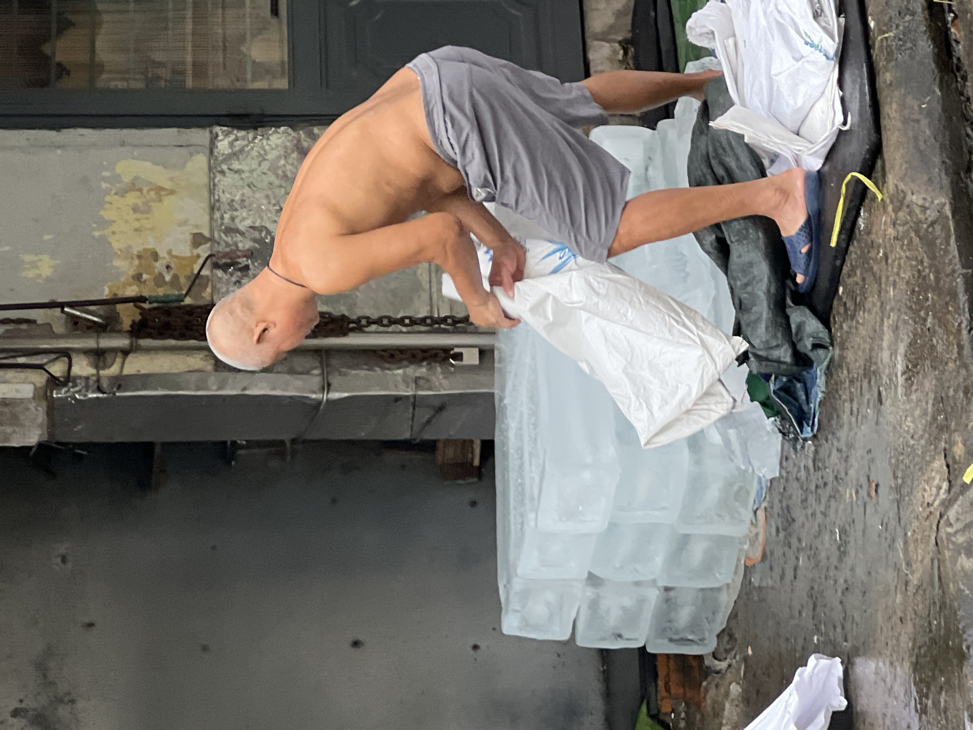 An unclothed man is seen from the side, handling a large white bag next to a stack of ice blocks. He is wearing gray shorts and blue sandals. The scene appears to be outdoors, with a wet, cobbled ground. Behind the man and ice blocks, there is a building with textured walls and a closed door. The image is likely from Nha Trang, Vietnam, suggesting a hot climate where ice is a necessity. The man's actions imply he is involved in the delivery or distribution of ice.