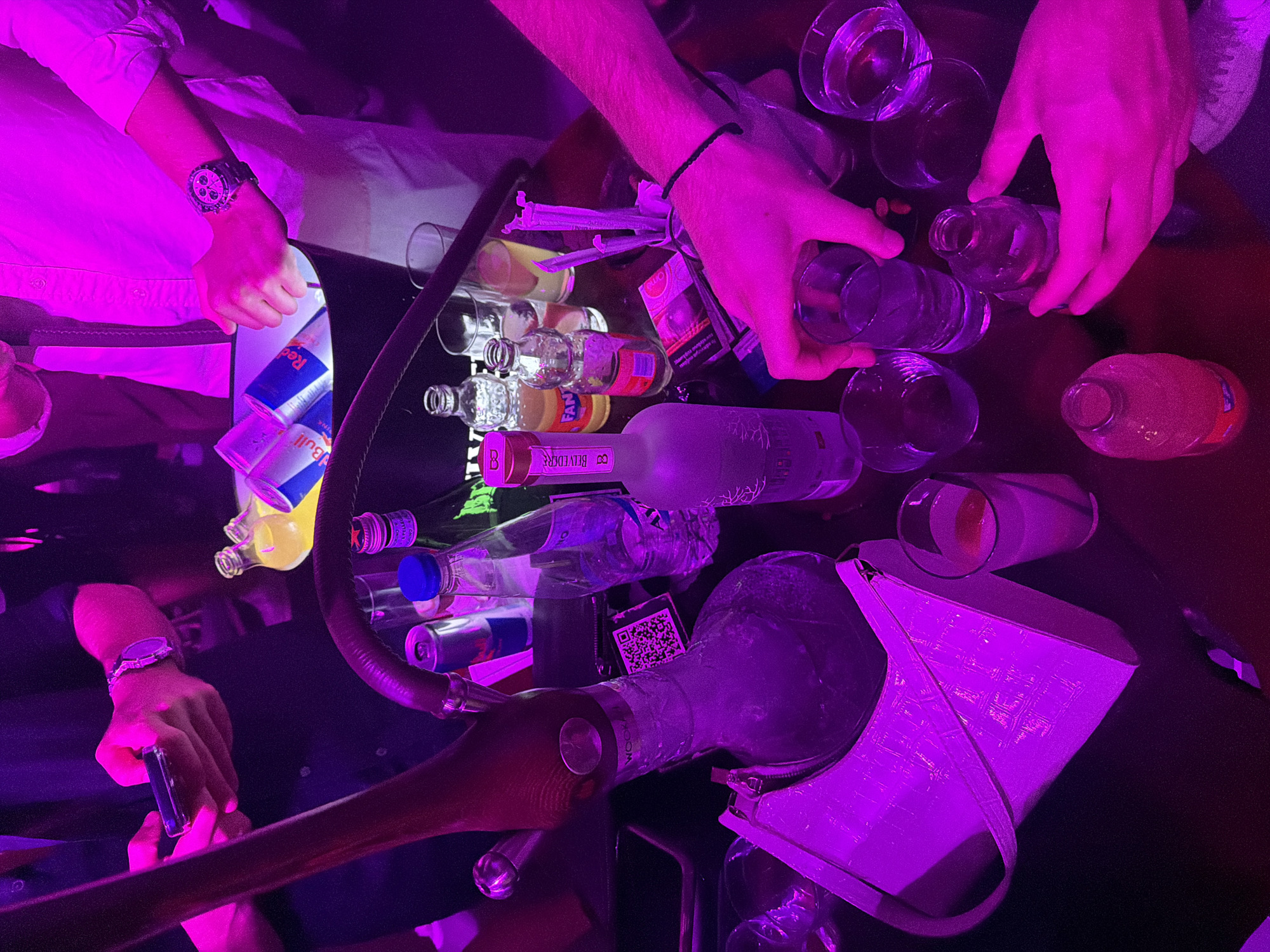 The image depicts a dimly lit indoor scene, likely a bar or club, with intense purple and pink lighting. The setting appears to be in the city of Kassandra Municipal Unit, Greece, though this is an assumption based on the provided context and cannot be definitively determined from the visual information alone.
In the foreground, hands are visible interacting with drinks and objects on a dark, reflective surface, likely a table. One person's hands are holding a glass and reaching for a bottle. Another person's hand is holding a smartphone. A hookah pipe with a visible stem and bowl is positioned to the left.
Scattered on the table are various items, including several bottles of alcohol, a bucket containing Red Bull cans, glasses, a white handbag, straws, and what appears to be a QR code sticker. The overall atmosphere suggests a social gathering or a night out.