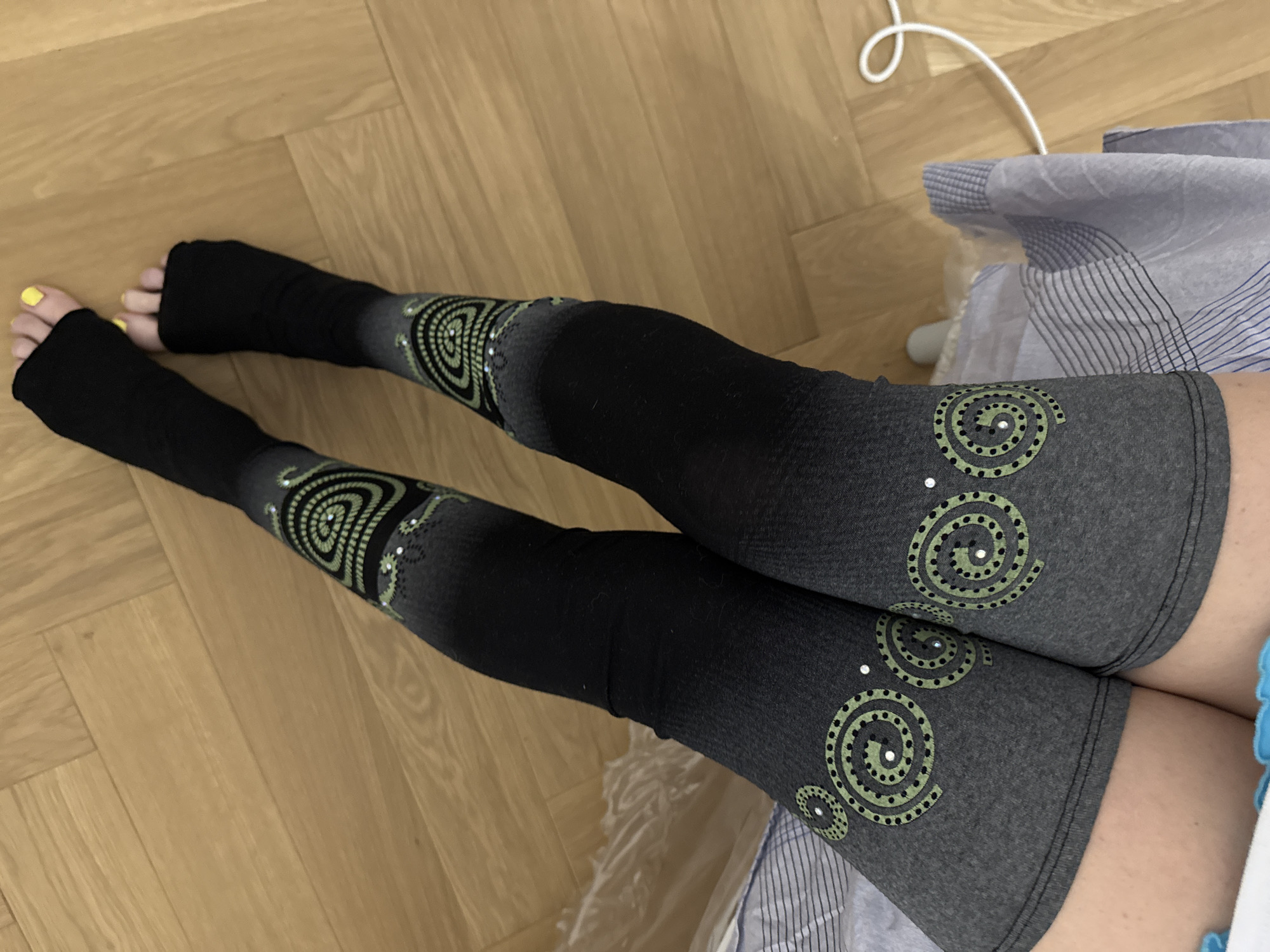 A high-angle, medium shot displays a person's legs clad in gray and black leggings adorned with circular patterns. The leggings have an open-toe design. The feet show toes painted yellow. The leggings transition from gray at the top, near the upper thighs, to black covering the feet. The legs are positioned on a light brown wooden floor. Part of a striped blue and white patterned fabric is visible to the right, near the upper legs. A white electrical cord snakes across the upper right corner of the floor. There is no indication of location of the city of Moscow, Russia.