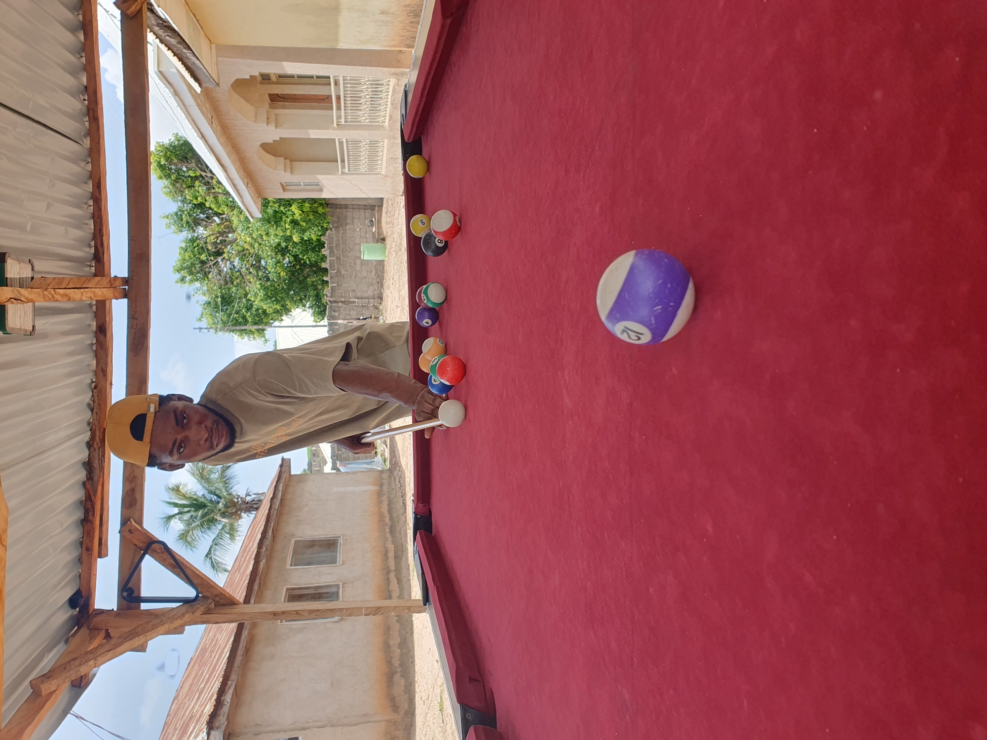 A young Black male wearing a tan t-shirt and a yellow baseball cap is playing billiards outdoors. He is positioned behind a red felt billiard table, holding a cue stick and aiming at a white cue ball. Several colored billiard balls are scattered on the table. The scene is set in a semi-rural environment, with buildings and vegetation visible in the background. A corrugated metal roof supported by wooden beams covers the immediate area of the billiard table, suggesting an outdoor recreational space. The buildings appear to be modest, possibly residential, with neutral-colored walls and tiled roofs. A palm tree can be seen in the middle ground. The sky is overcast.